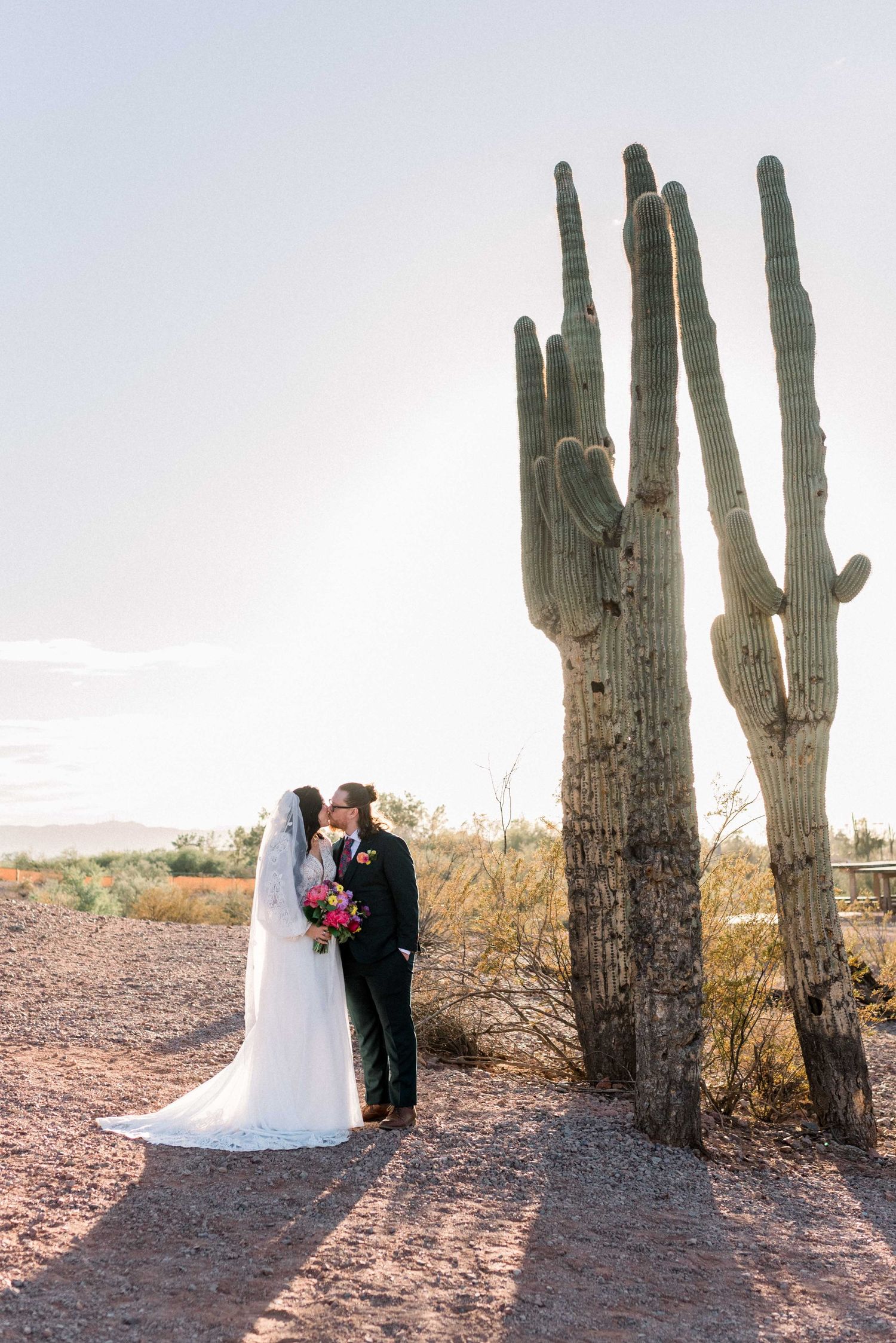 A couple poses together in a desert landscape with tall saguaro cacti silhouetted against a bright sky.