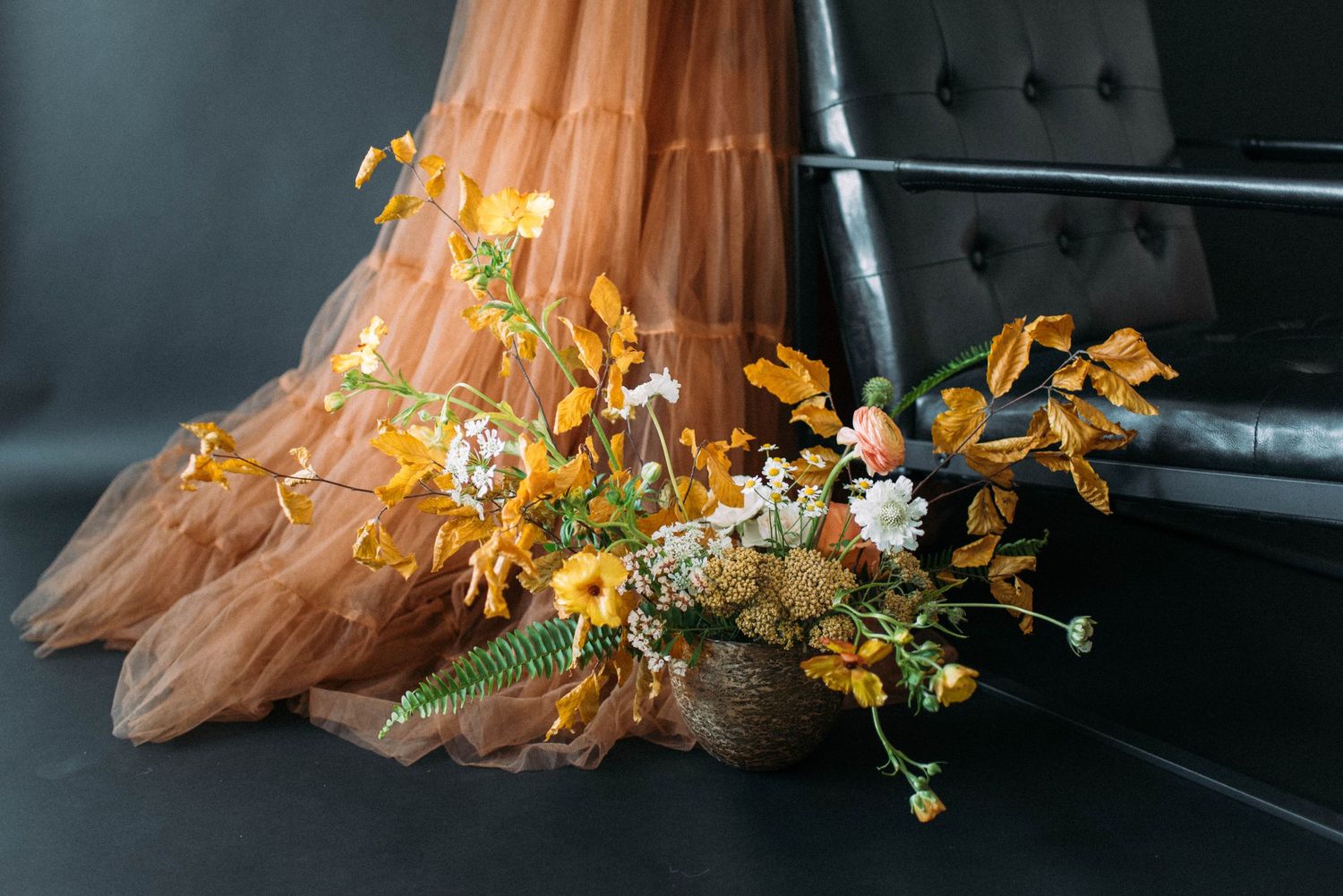 A floral arrangement with golden leaves rests beside flowing brown tulle fabric.
