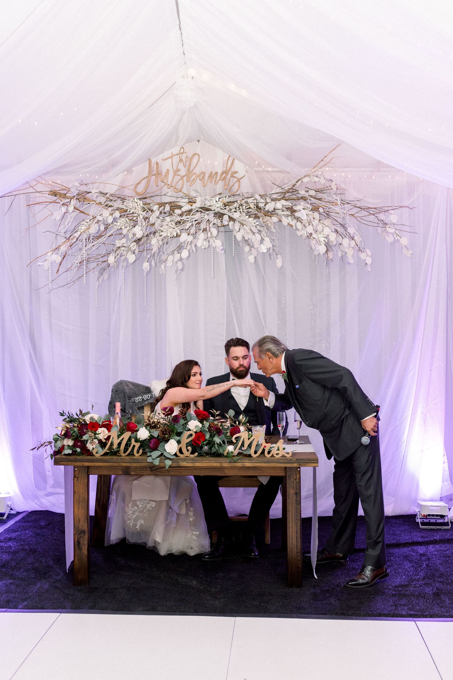 Wedding couple seated at a decorated sweetheart table under a floral chandelier in a draped white tent.