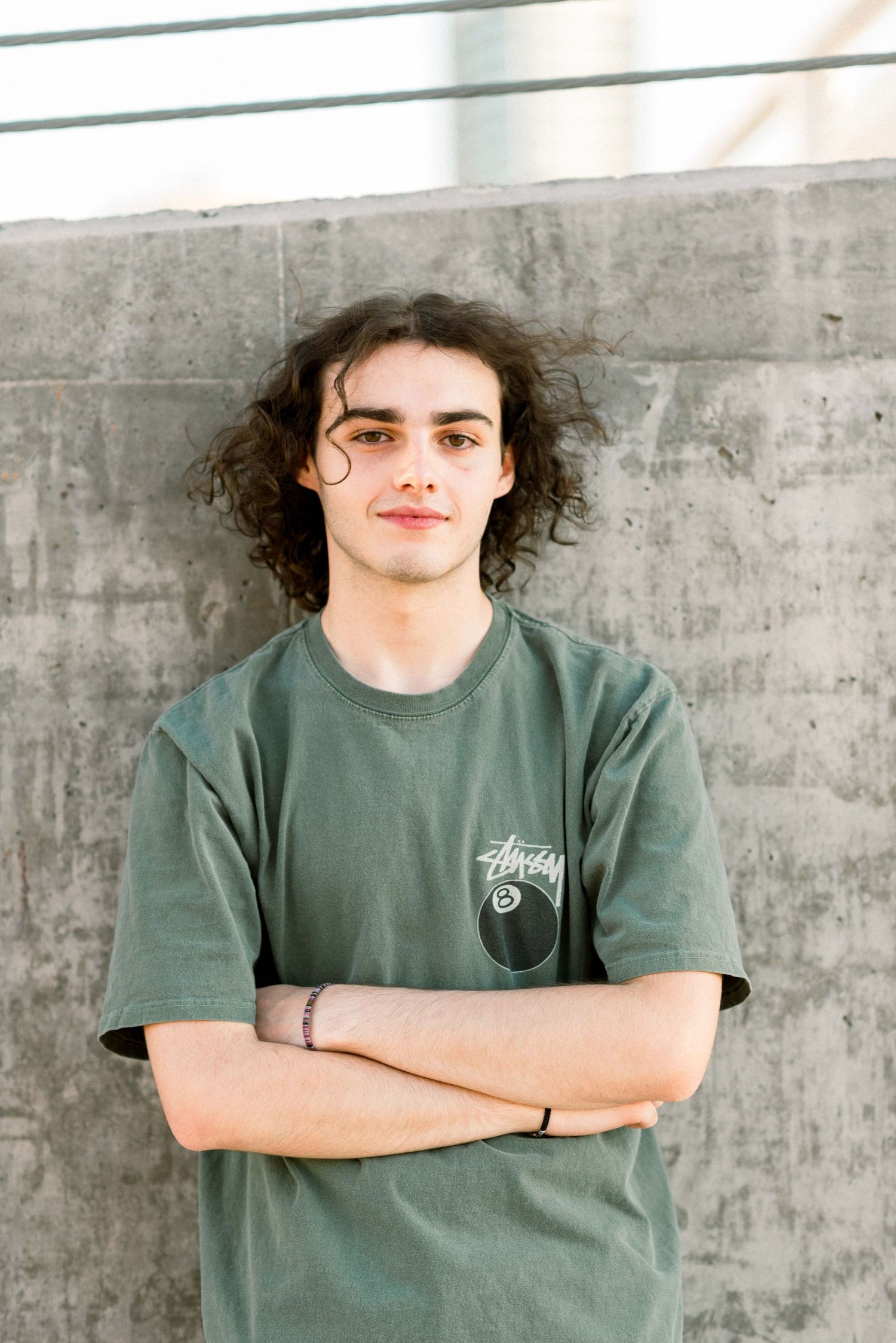 Person wearing green Stussy t-shirt stands with arms crossed against concrete background.