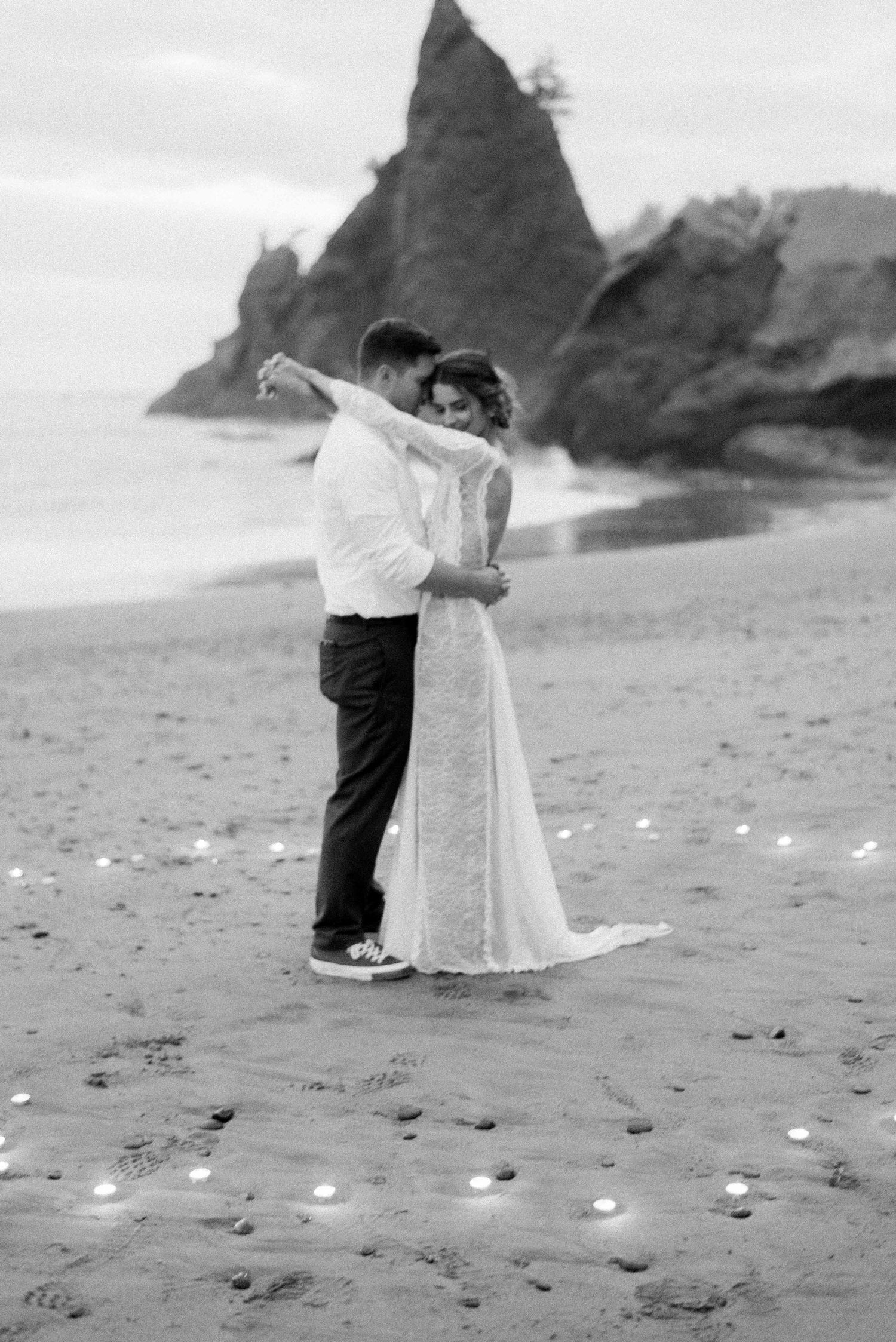 A romantic black and white photo of newlyweds embracing on a beach with dramatic rock formations.