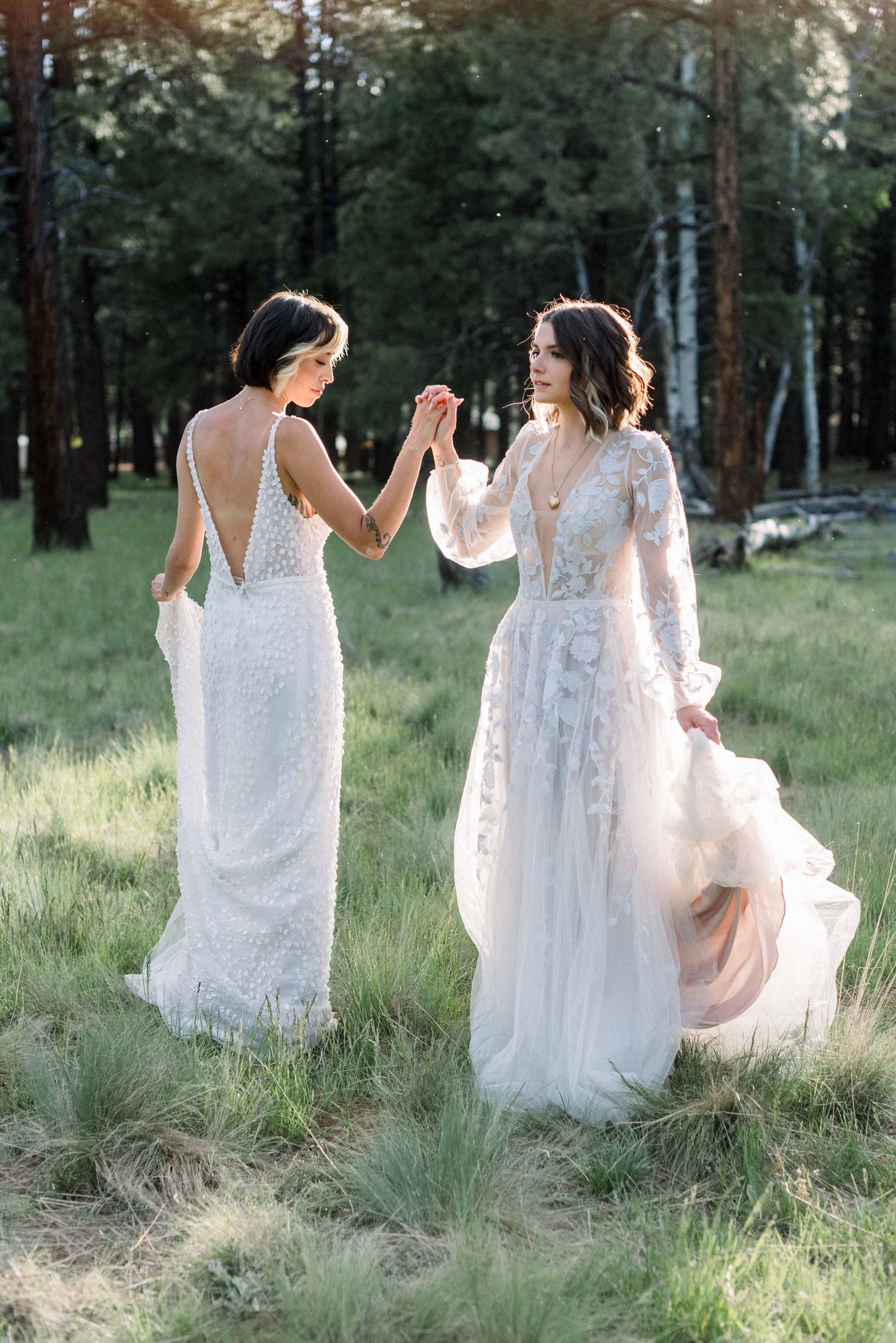 Two people in white wedding gowns hold hands while standing in a sunlit forest clearing.
