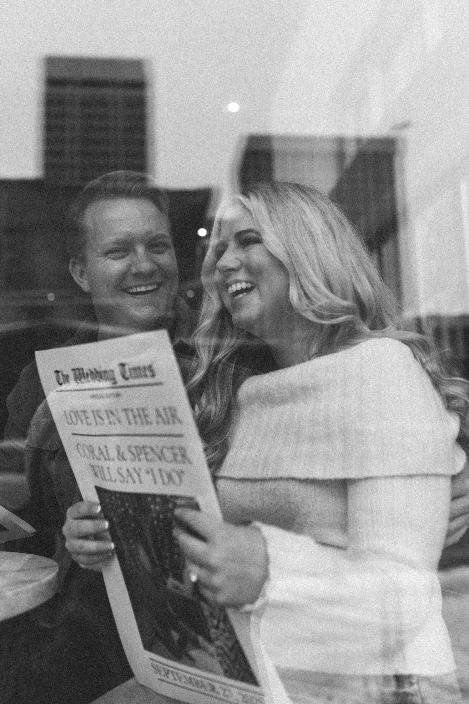 Black and white photo of a couple laughing while reading a custom newspaper at a cafe.