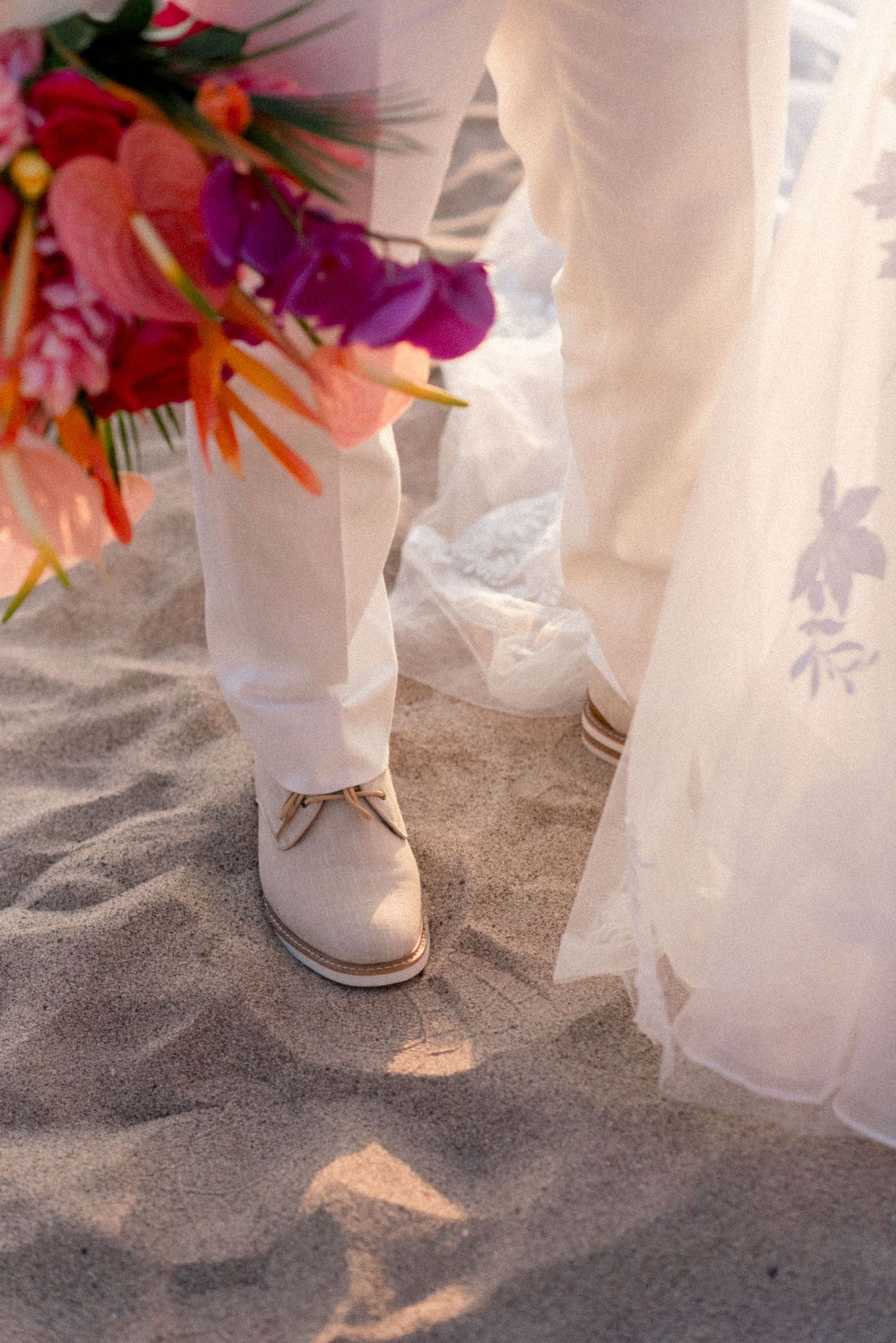 White dress and suit details with colorful tropical bouquet on sandy beach.