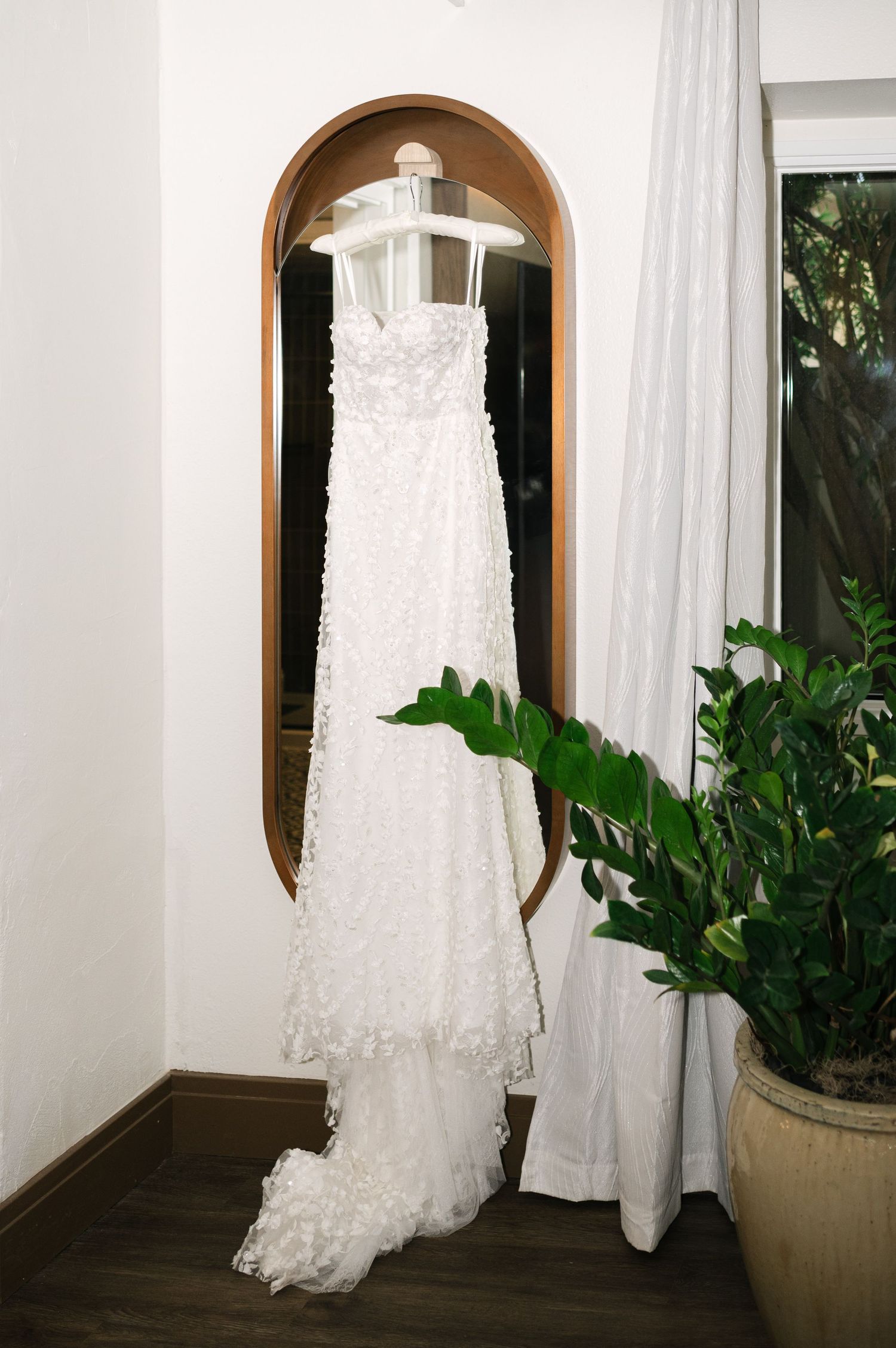 Elegant lace wedding dress hangs by arched windows with white curtains and indoor plants.