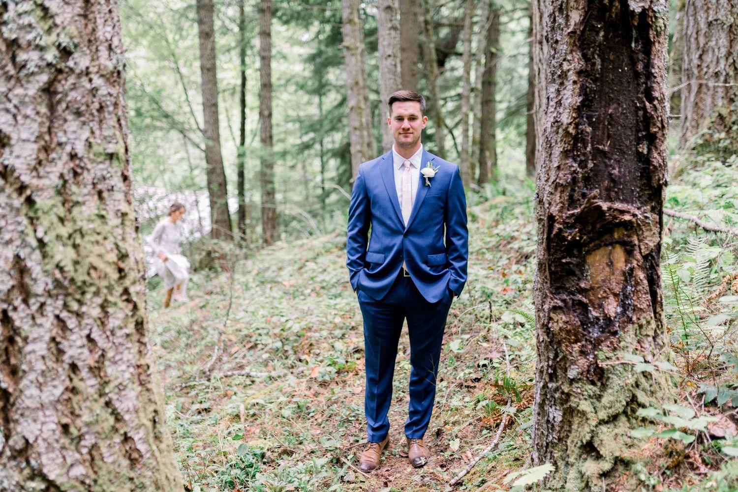 Groom in blue suit standing alone among tall trees in a forest setting.