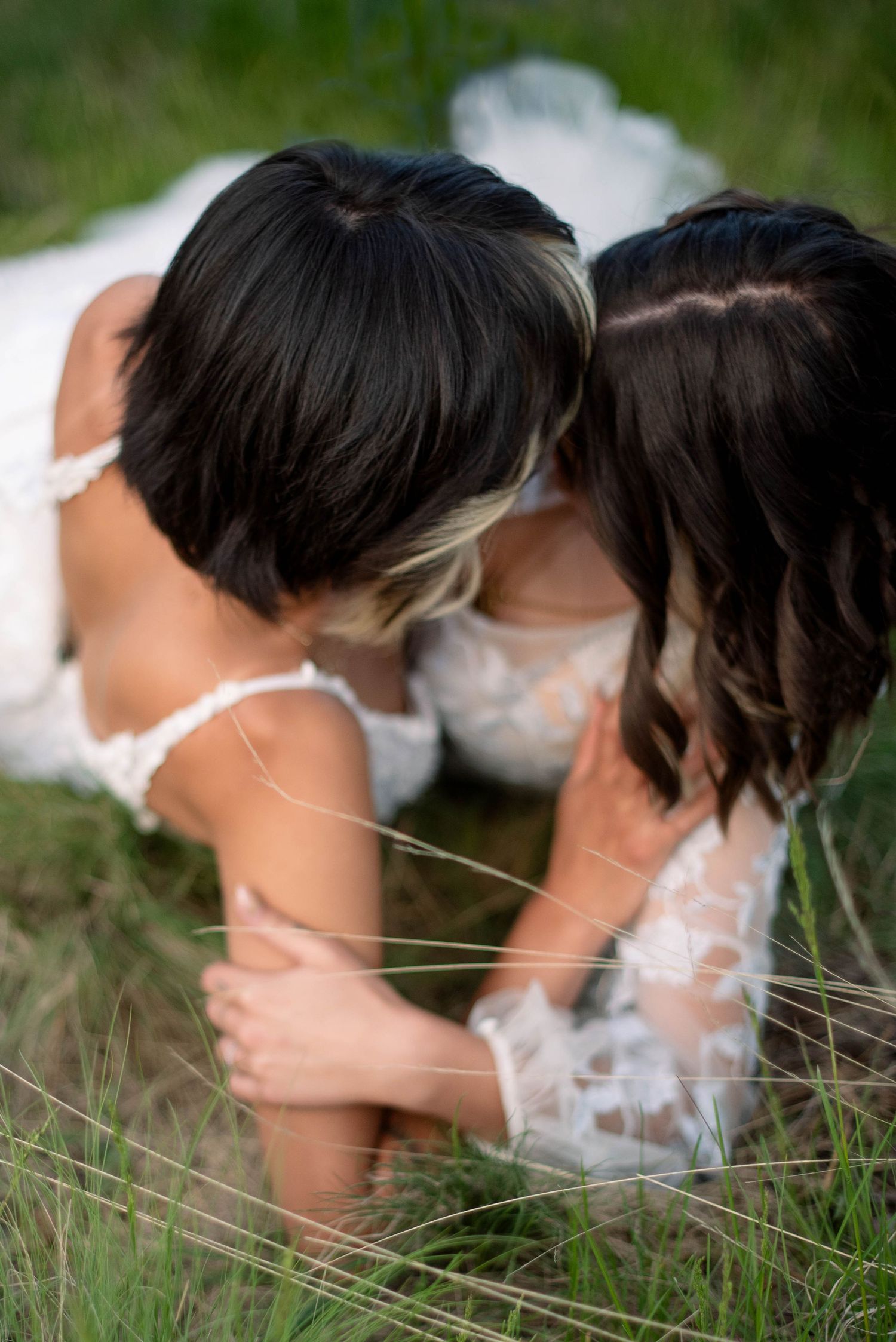Two people in white dresses laying together in tall grass during an outdoor photoshoot.