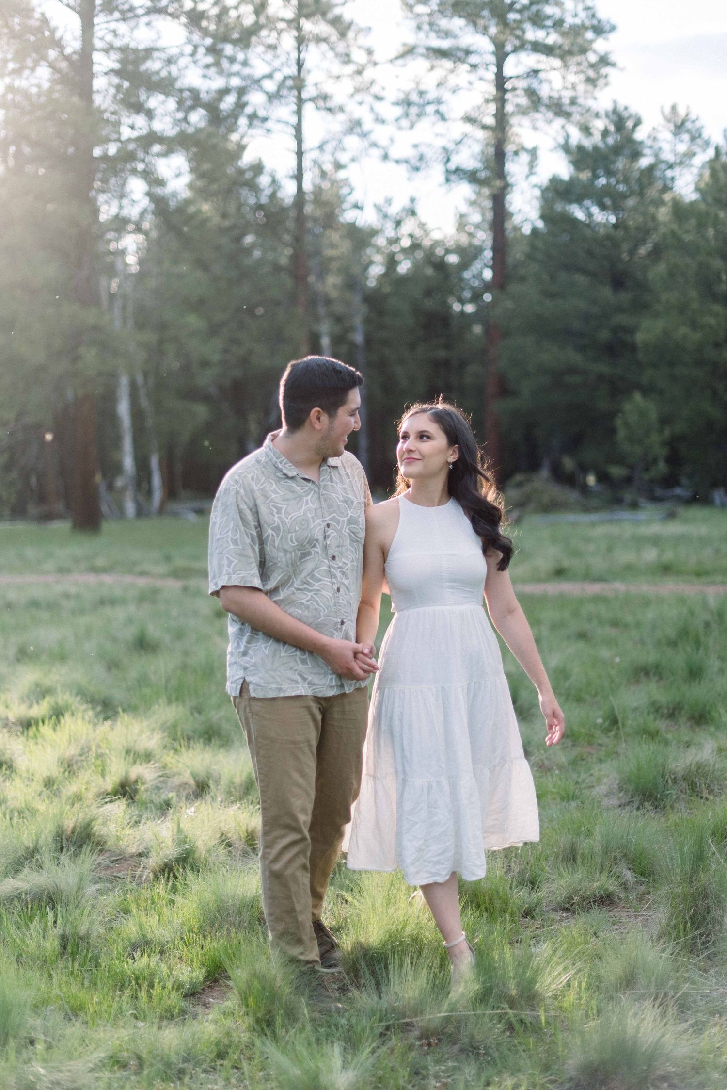 A couple strolls together through a grassy meadow with pine trees in the background.