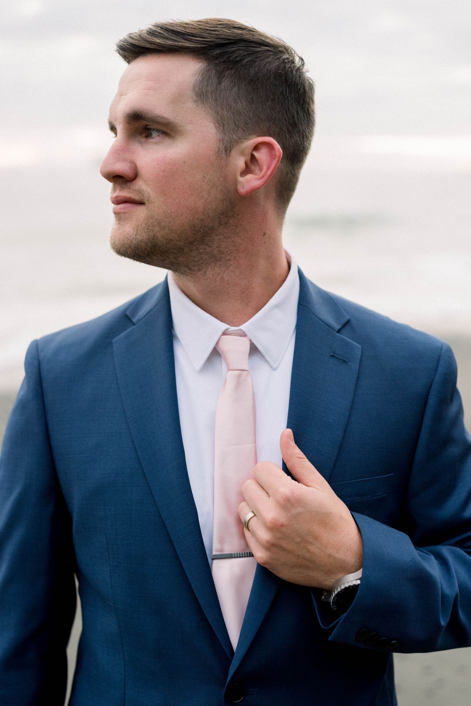 Side profile portrait of person in blue suit with pink tie against ocean backdrop.