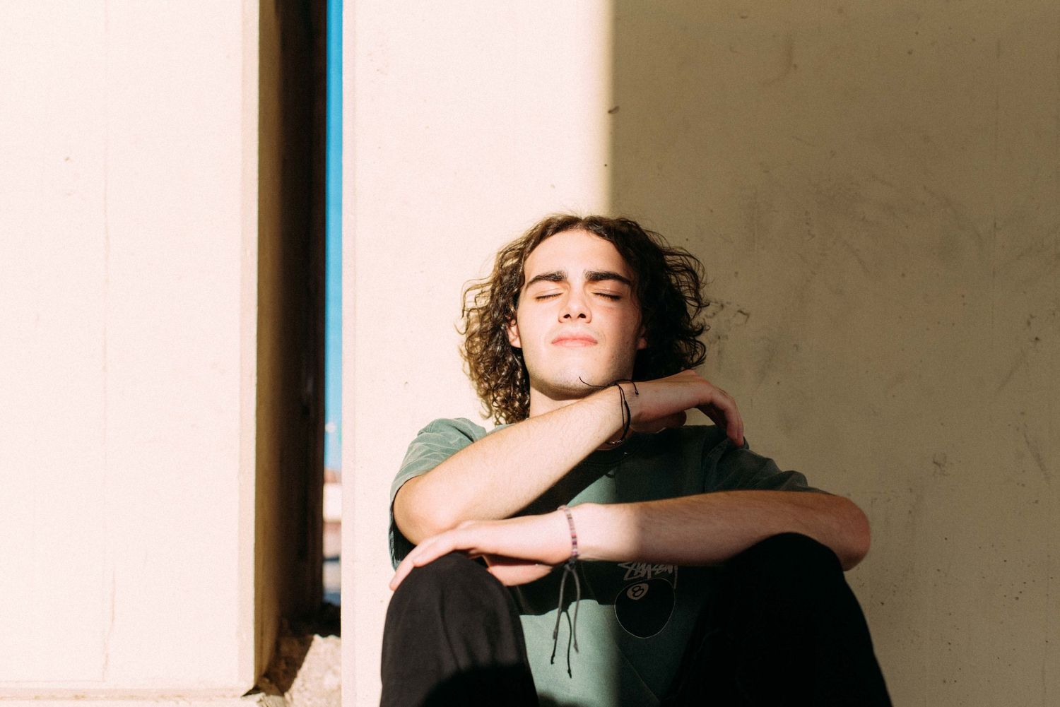 Candid sunlit portrait of person in green shirt resting against wall.