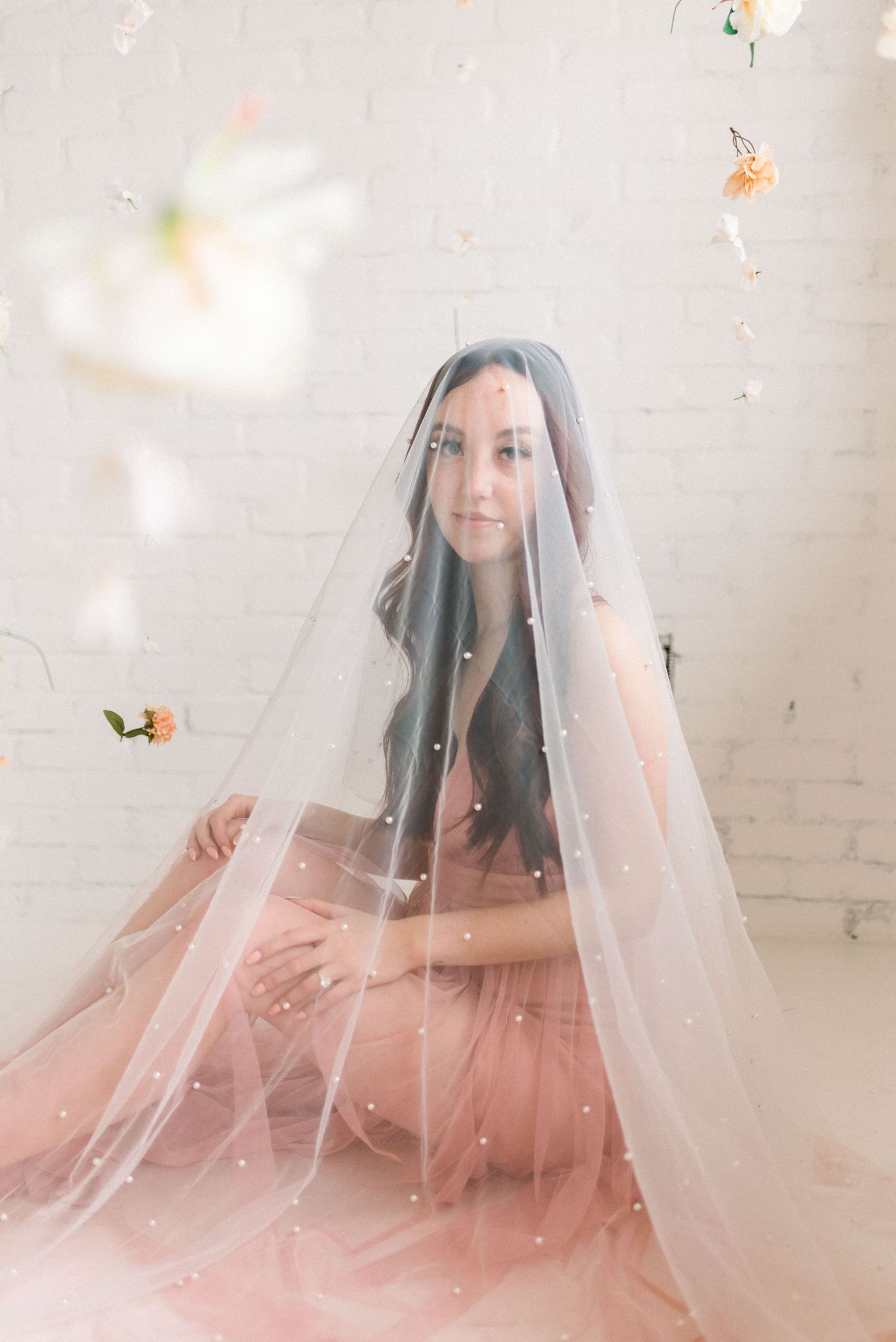 Ethereal portrait under sheer veil with floating floral elements and pink gown.