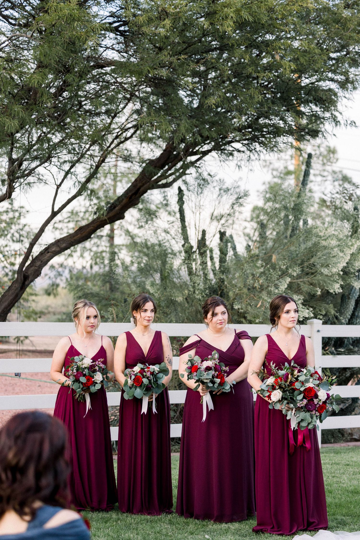 Four bridesmaids in burgundy gowns hold floral bouquets by a white fence.