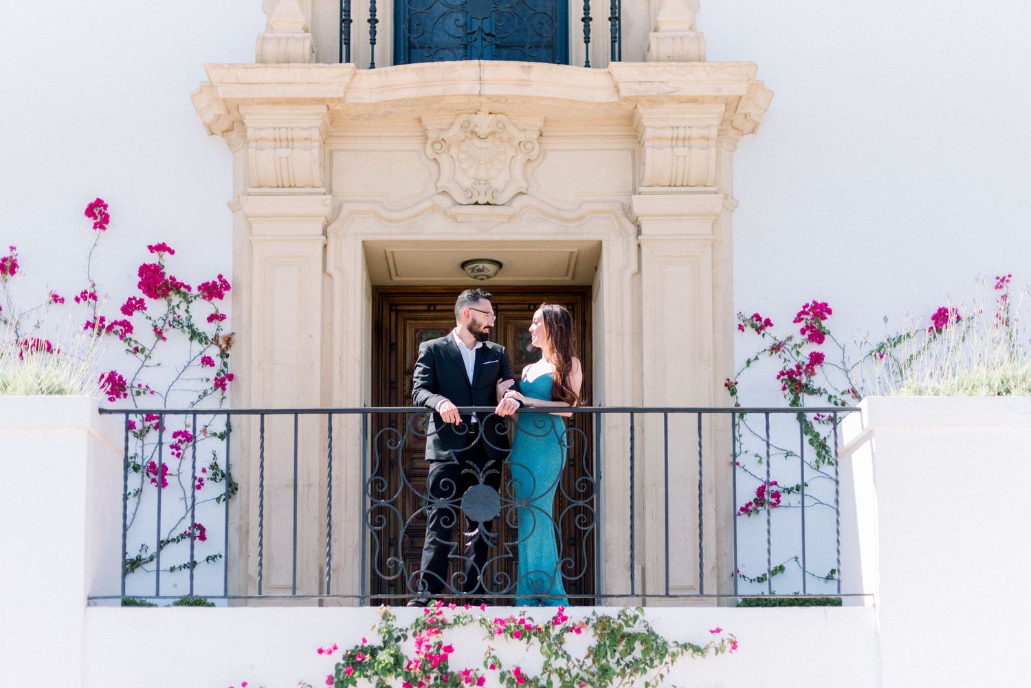 Couple poses on balcony of white Mediterranean building with pink bougainvillea flowers.