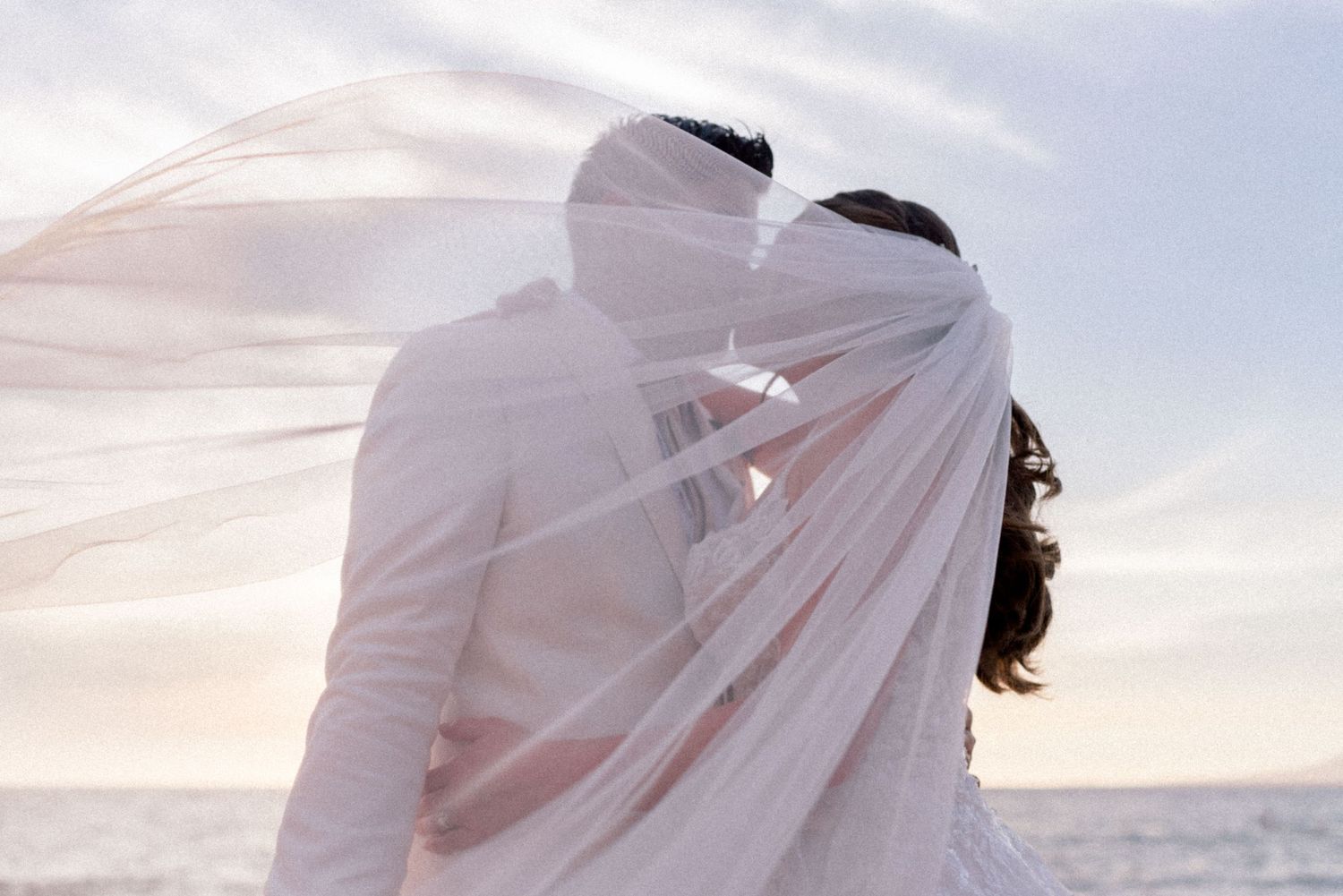 Romantic couple under flowing veil at beach during sunset.