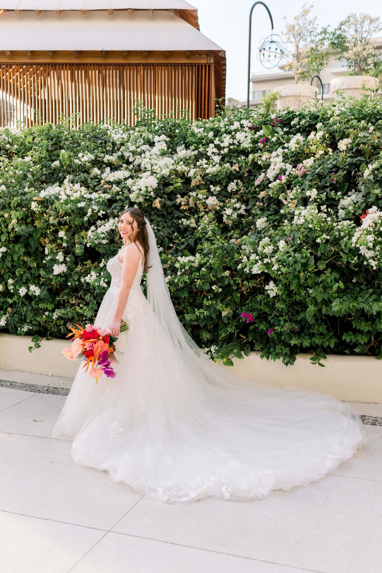 Bride in flowing ball gown poses against wall of white climbing roses with tropical bouquet.