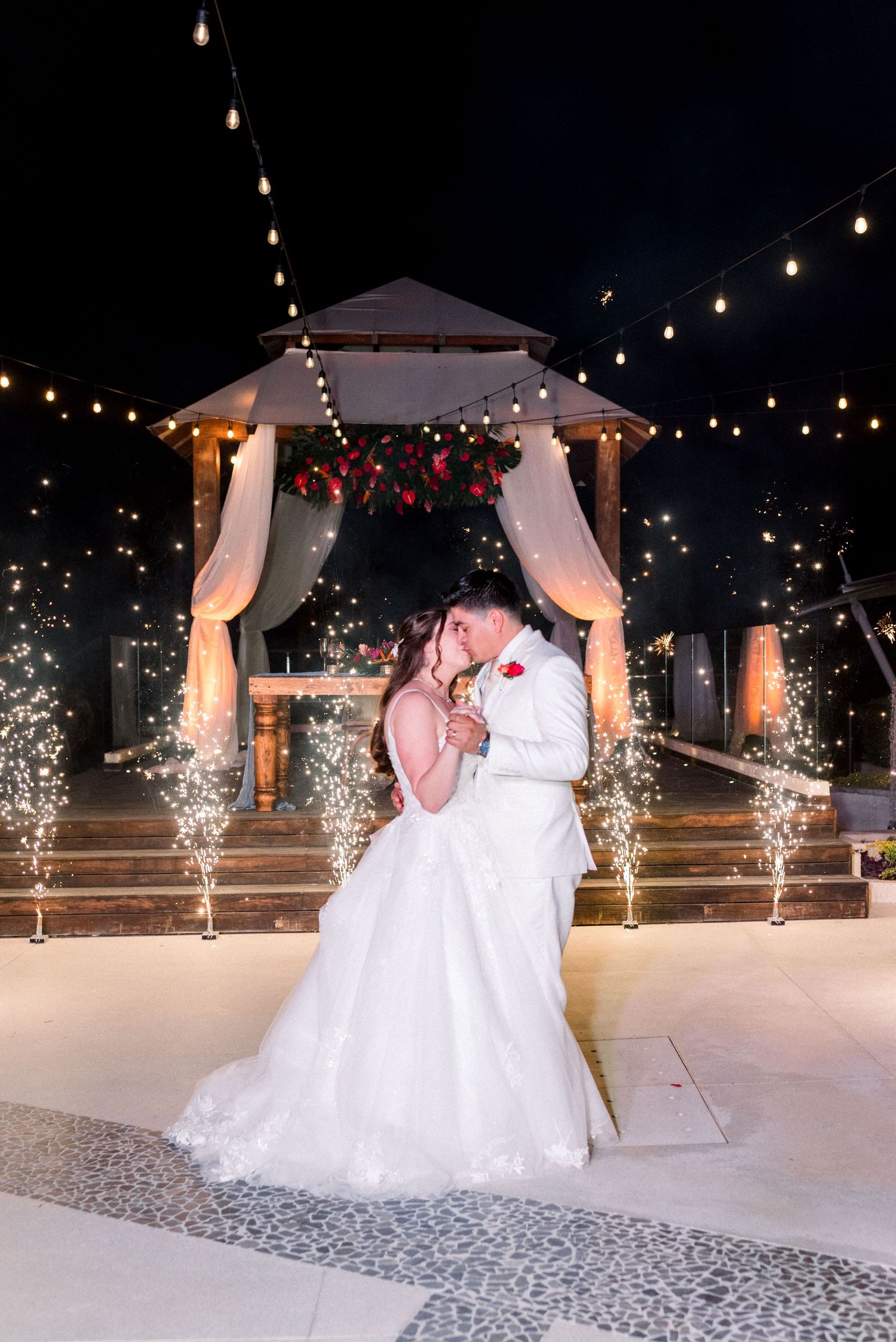 Newlyweds embrace during their reception under a decorated gazebo with sparkling fountains and twinkle lights.