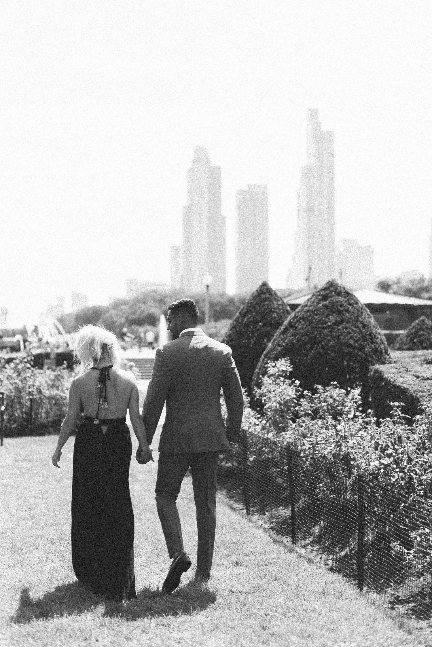 Black and white photo of couple walking through a garden with city skyline in distance.