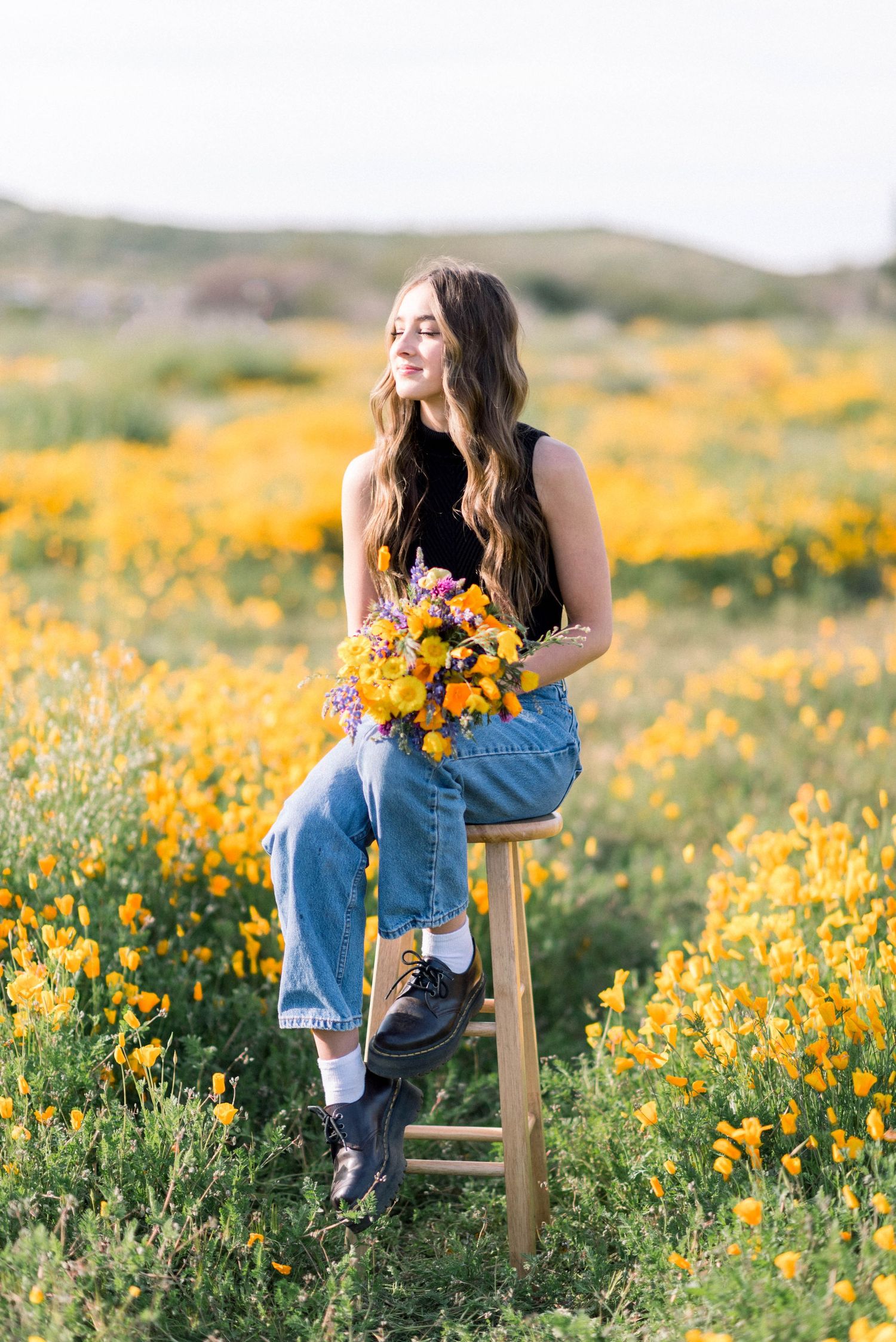 Person holding a vibrant yellow wildflower bouquet while seated on a stool in a meadow.
