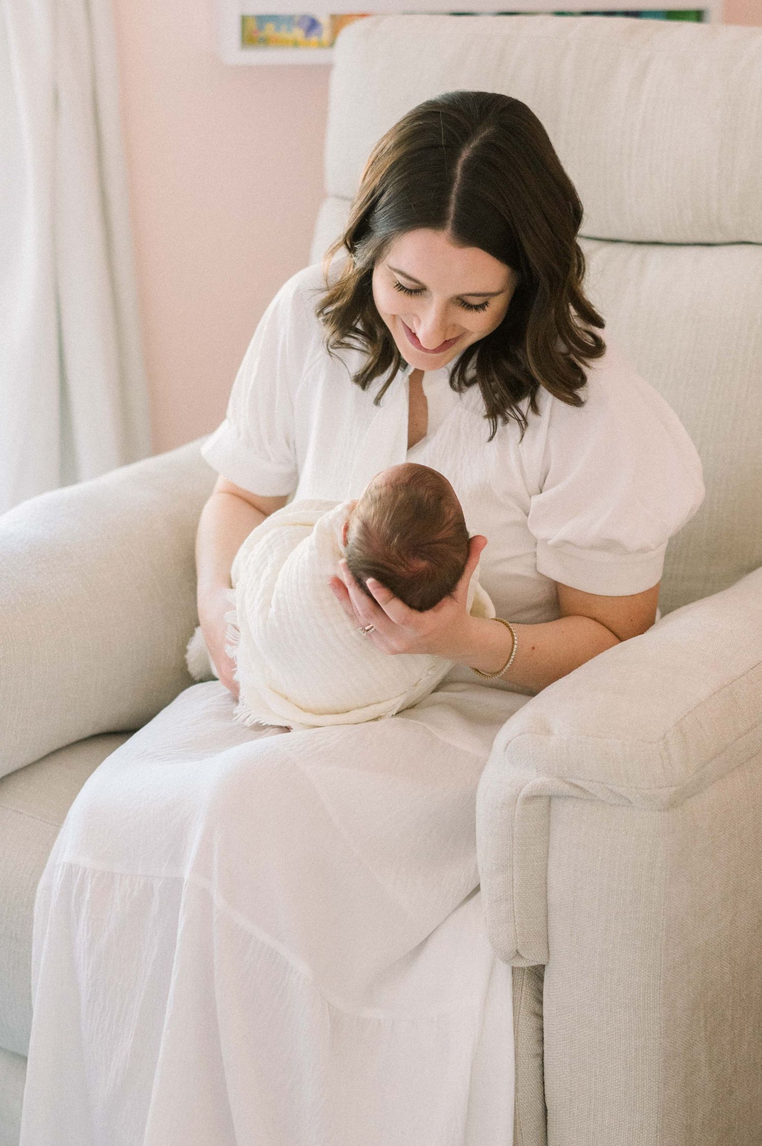 A tender moment as a parent in white cradles a swaddled newborn baby in a cozy armchair.