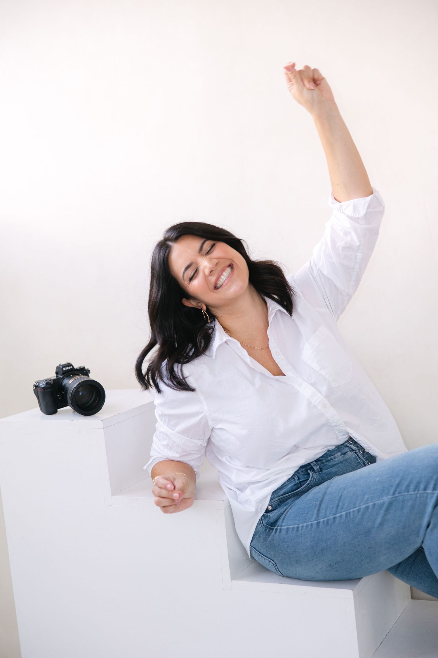 A person in a white button-up shirt and jeans poses playfully against a white wall in a photo series.
