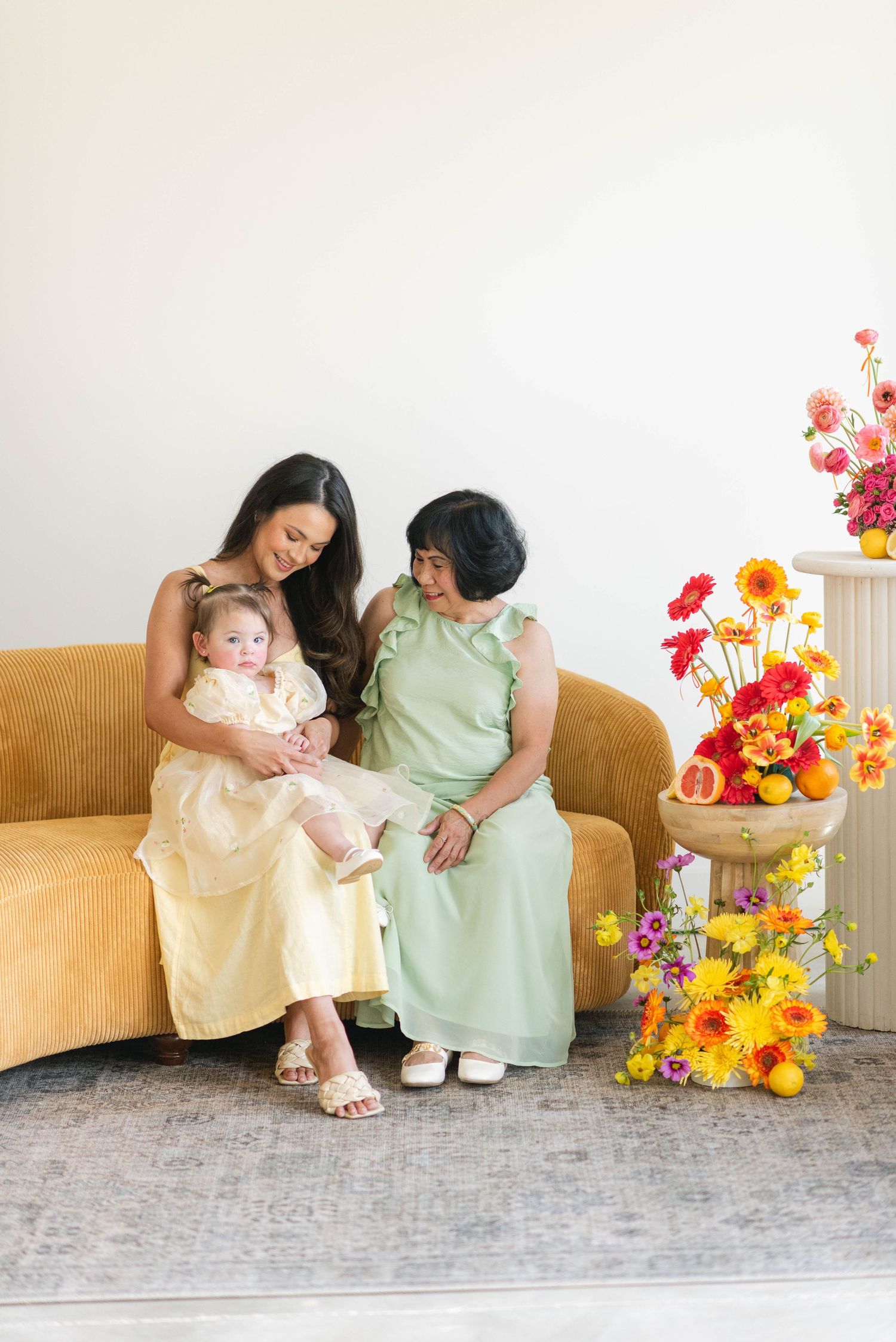 Two people sit together on a tan couch with vibrant orange and yellow floral arrangements in a bright studio setting.