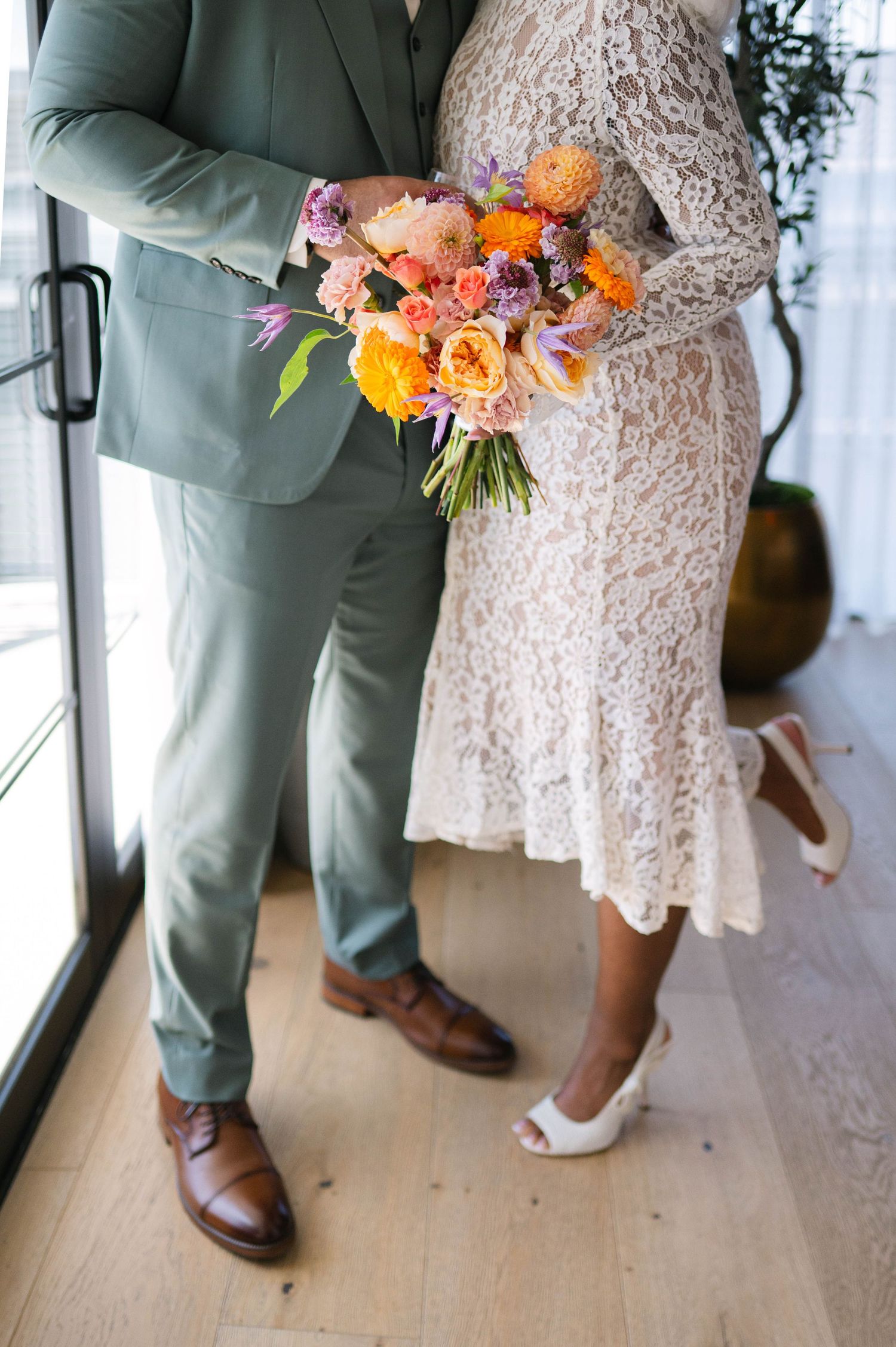 Wedding couple's feet and colorful floral bouquet with orange and pink blooms against a light wooden floor.