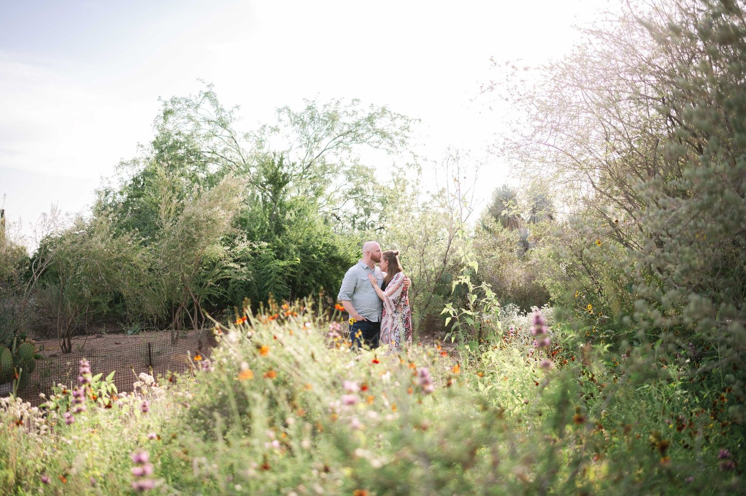 A couple walks through a lush garden path surrounded by blooming wildflowers and greenery in soft sunlight.