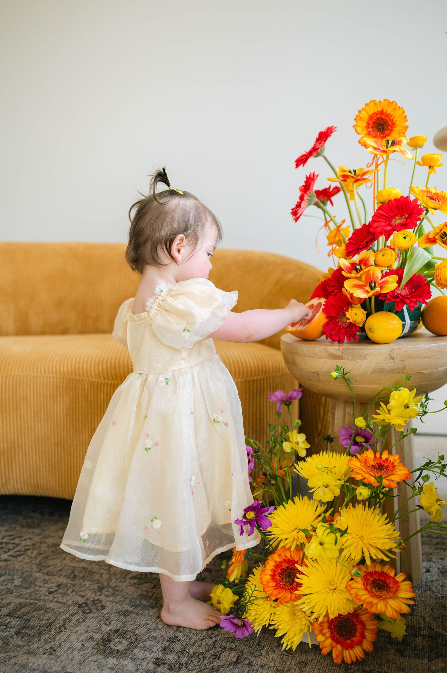 A small child in a white dress curiously explores a vibrant floral arrangement of sunflowers and daisies on a brown couch.