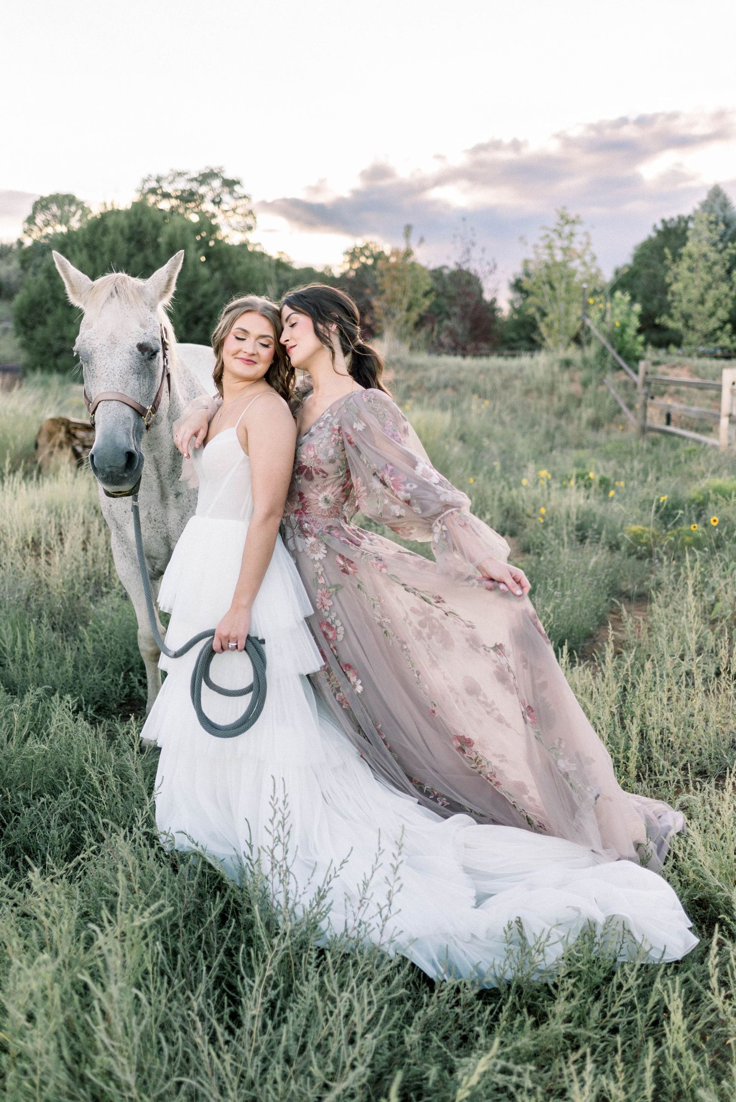 Two women in romantic dresses pose with white horse in sunlit meadow at sunset.