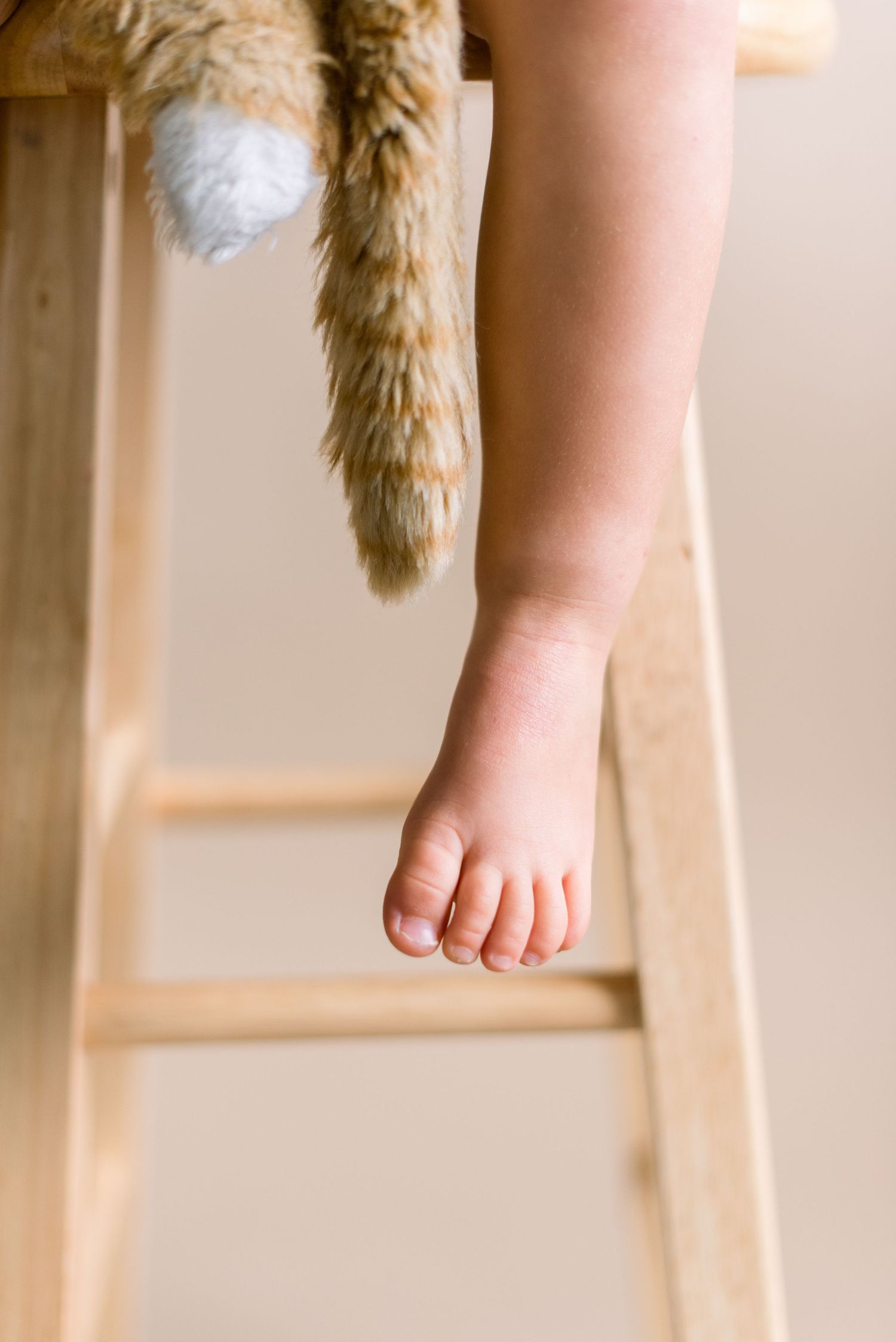 A tiny bare foot dangling next to plush toy tail on wooden stool.