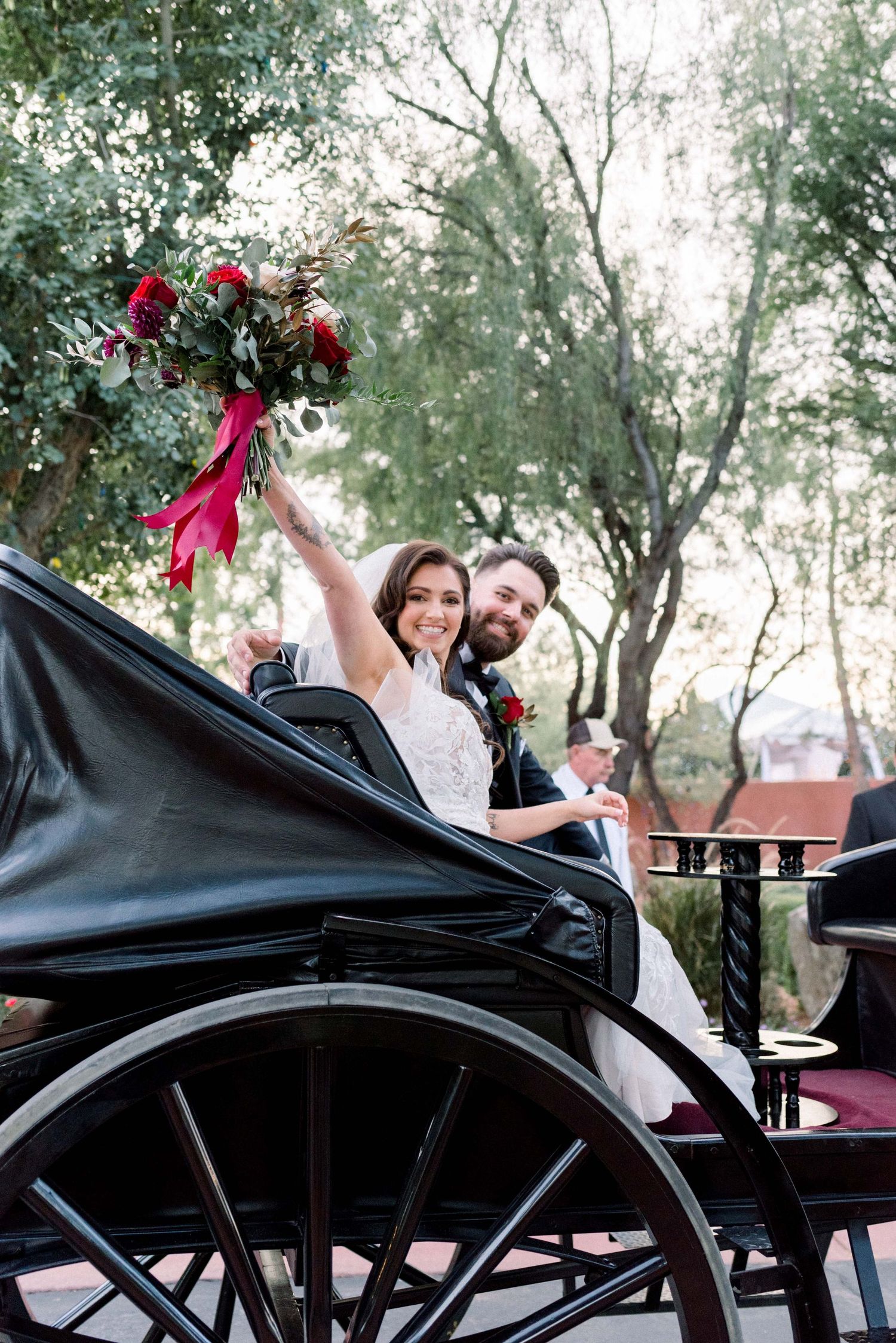 Couple celebrating in a vintage black carriage with red floral bouquet.