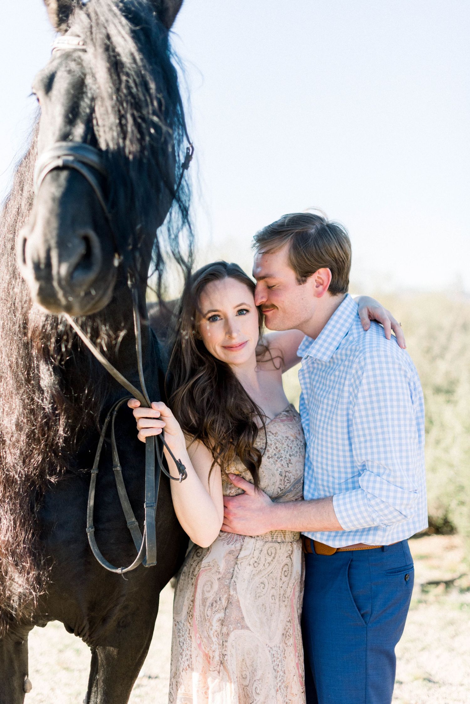 Romantic couple shares intimate moment beside black horse in desert setting.