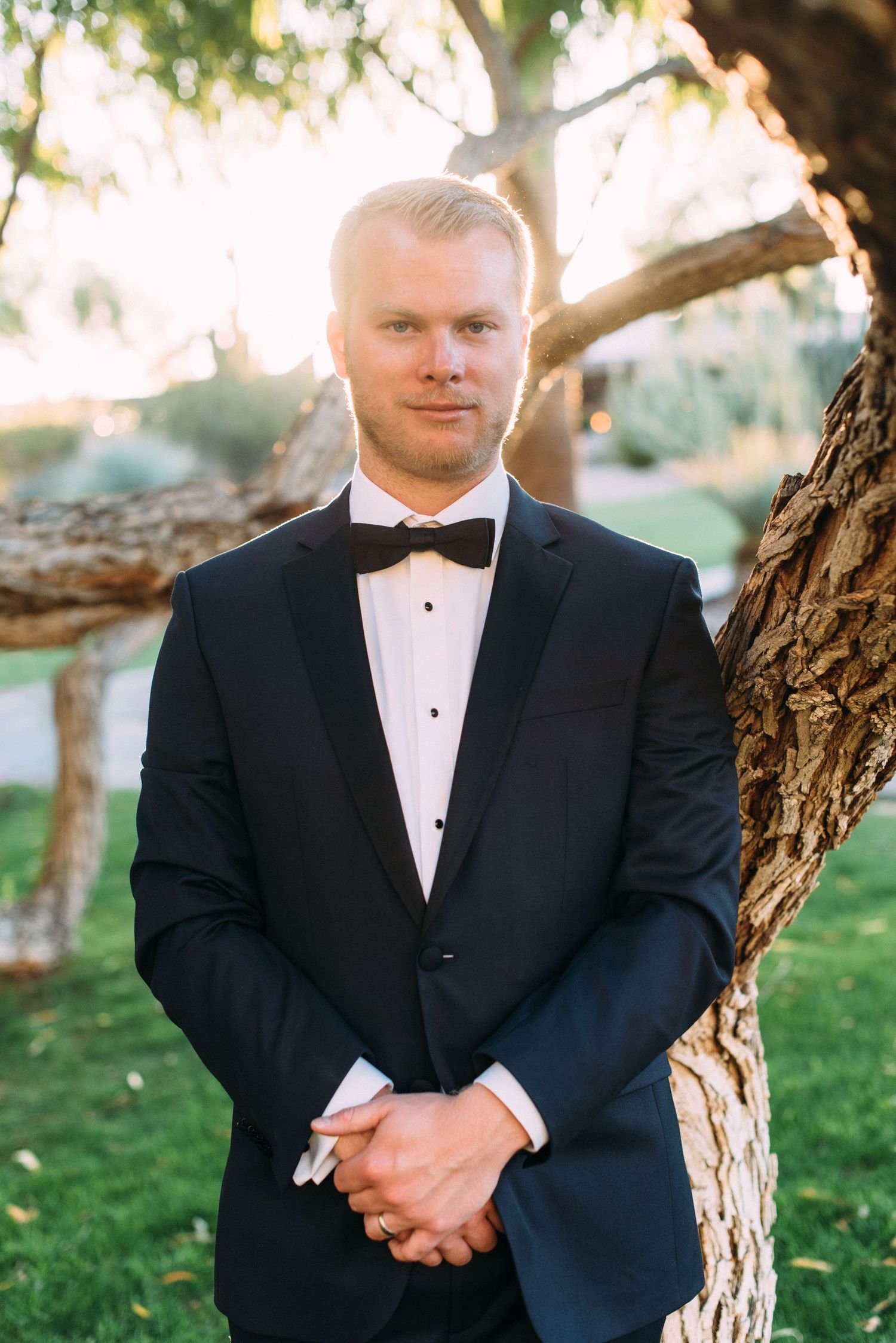 A formally dressed groom in a black tuxedo and bowtie stands against a natural outdoor backdrop.