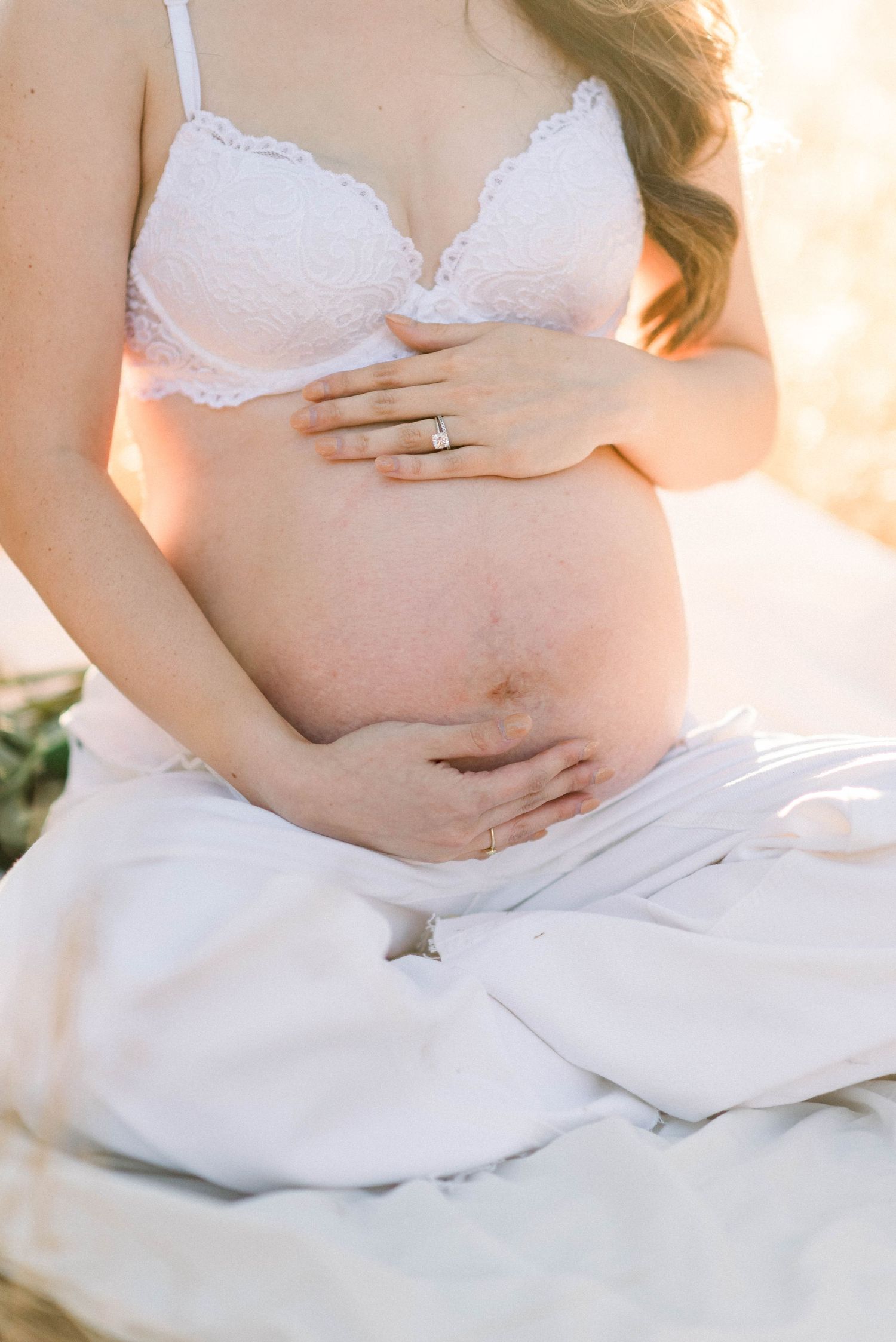 Close-up maternity shot showing pregnant belly with lace bra and wedding ring in soft light.