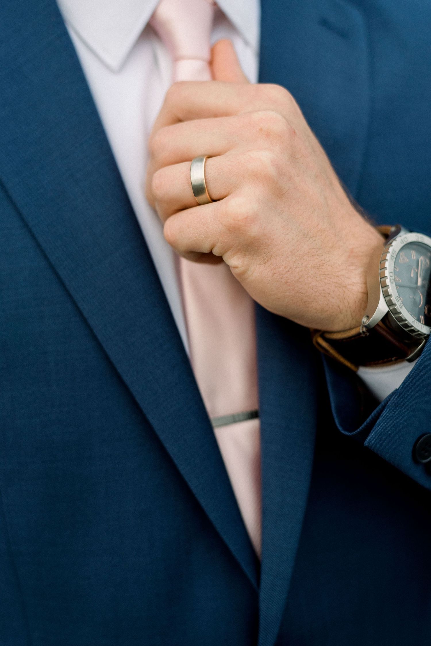 Close-up of a hand adjusting a tie showing a wedding ring and watch with a navy suit.