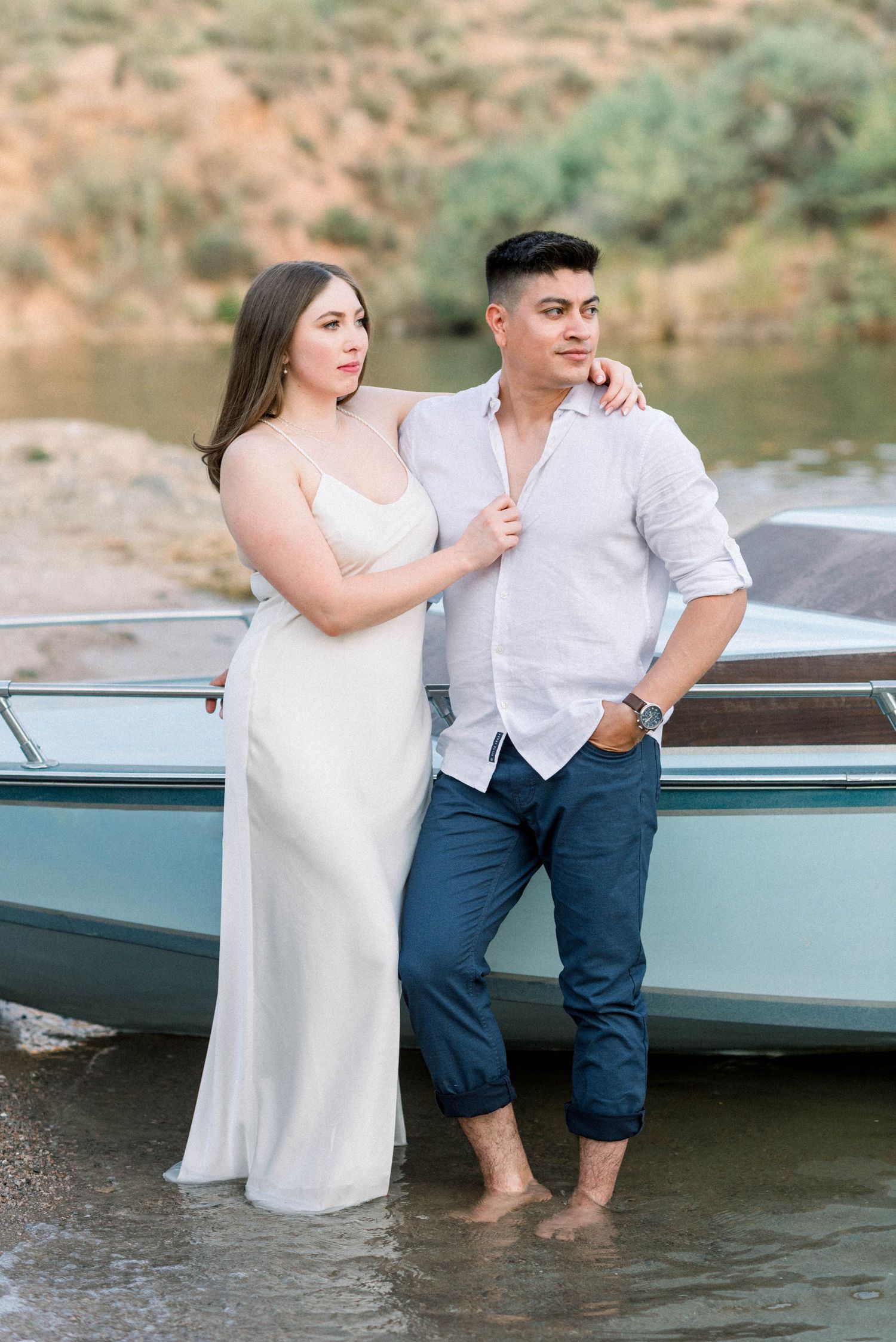 A couple poses together by a boat at the edge of a lake with rocky cliffs in the background.