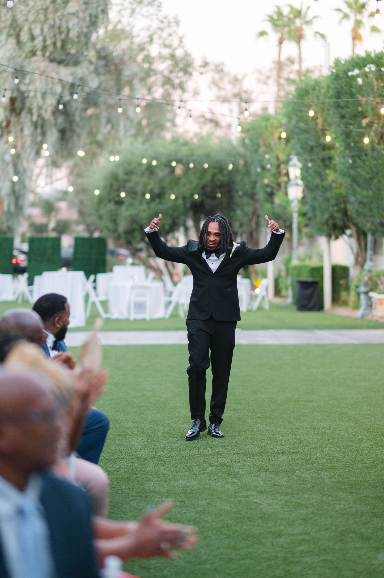 Person in black attire joyfully poses with arms spread wide on a grassy lawn with palm trees in the background.