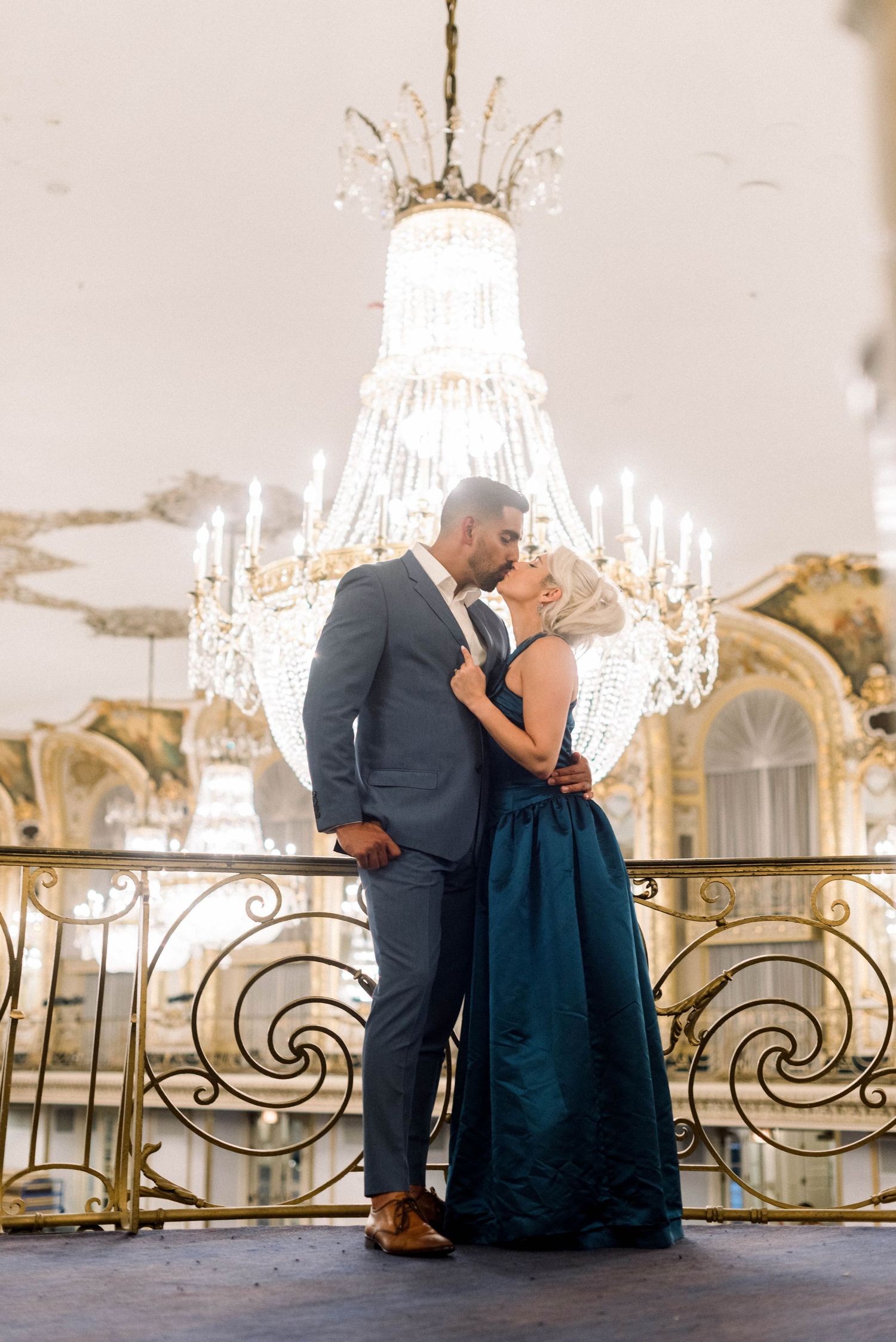 A romantic couple shares an intimate moment beneath an ornate crystal chandelier in a luxurious ballroom.