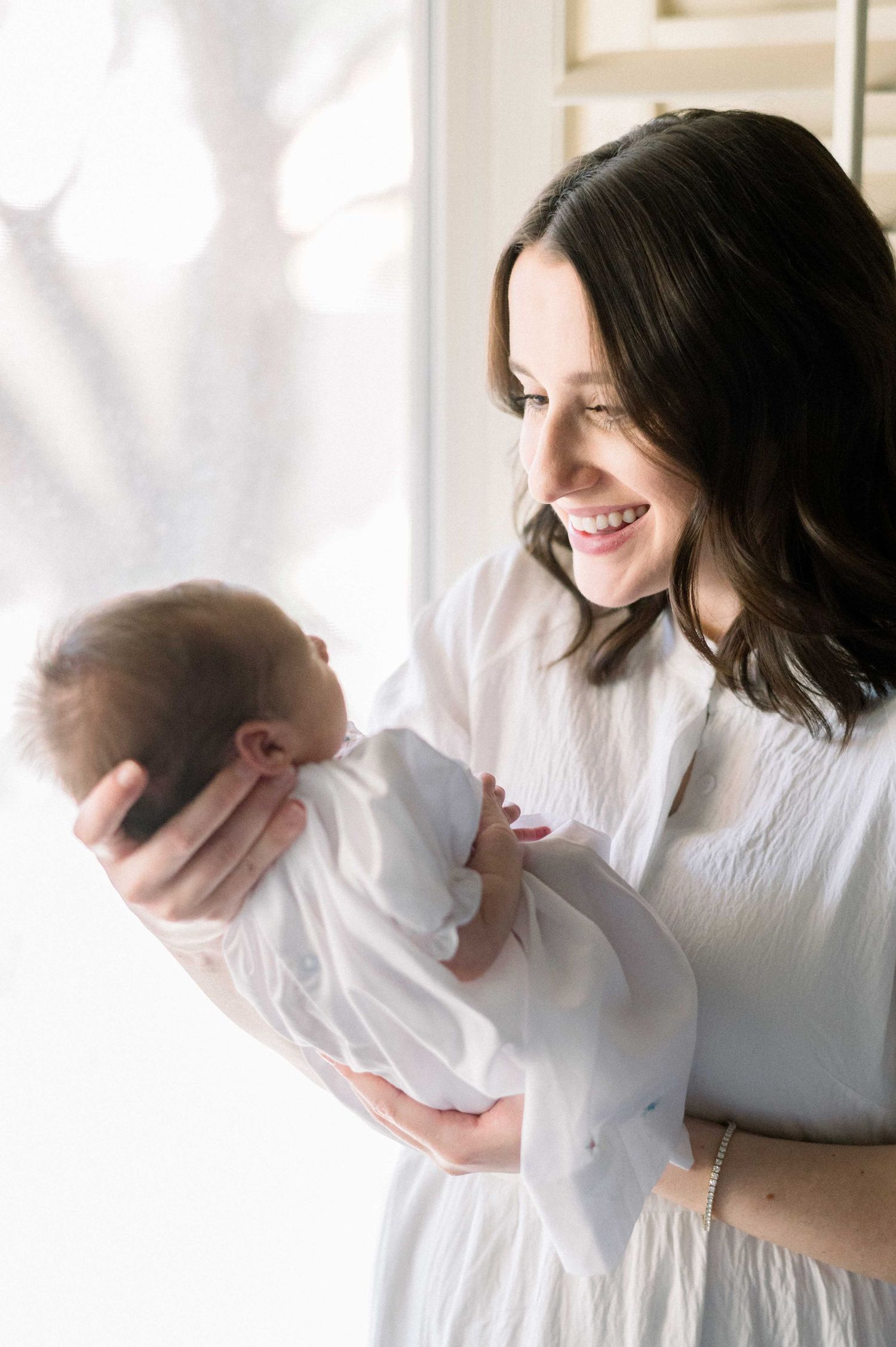 A woman in white holds a sleeping newborn baby near a sunlit window.