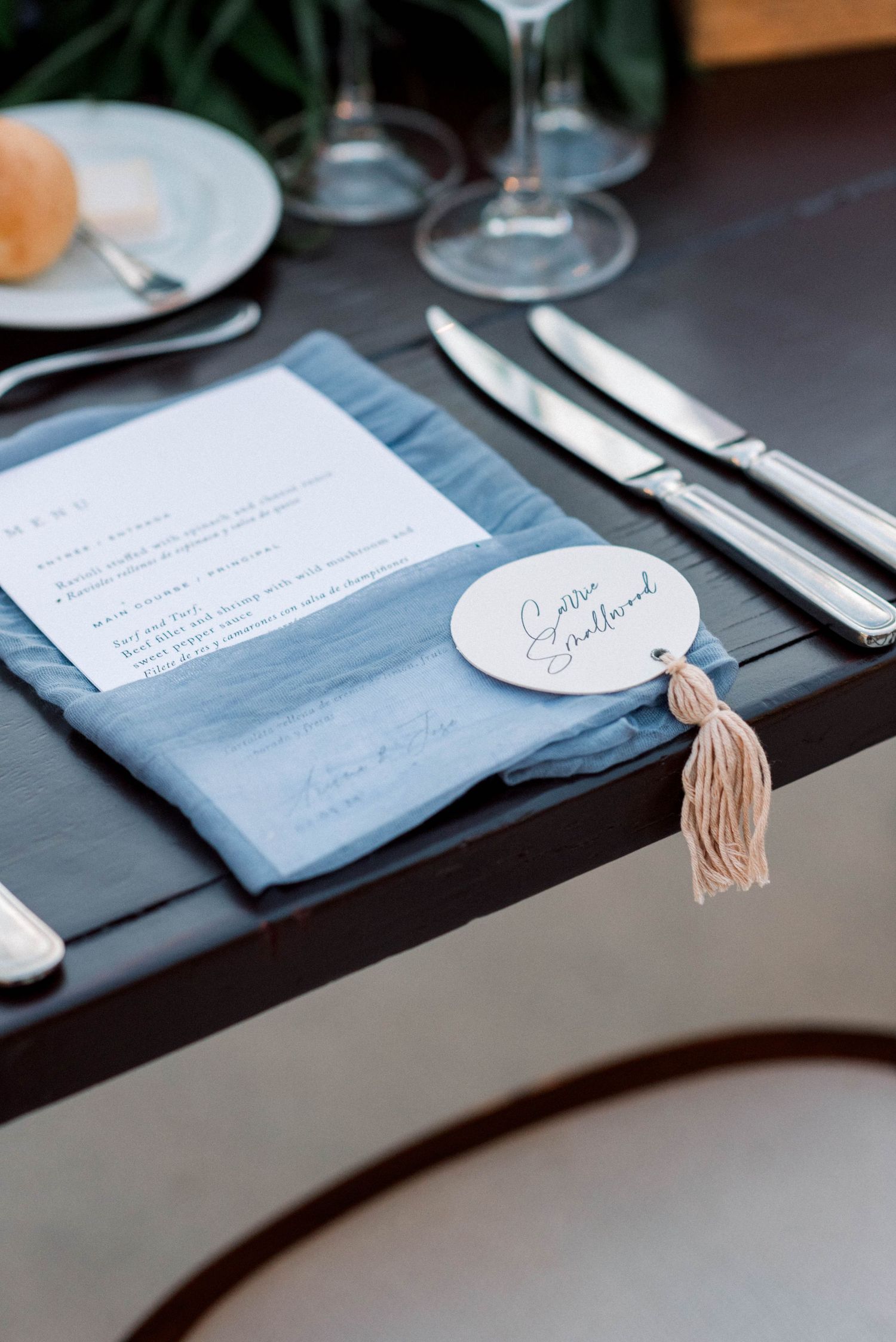 Wedding place setting with blue napkin, tassel detail, and elegant dinnerware on dark table.