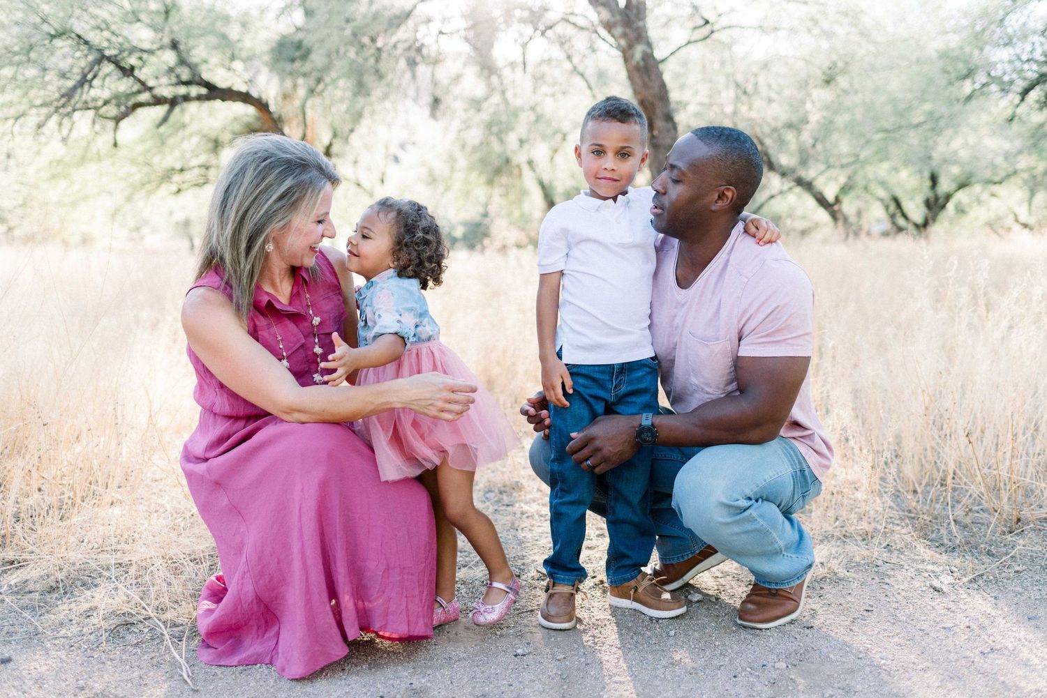 Family sitting together and smiling on nature trail with tall grass and trees.