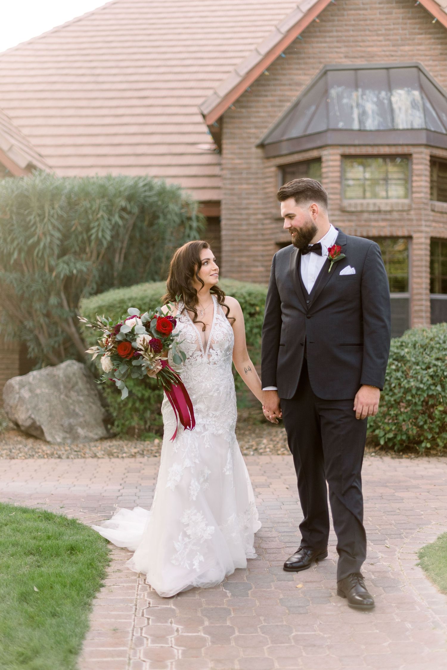 Bride in lace gown and groom share tender moment on stone pathway outside brick venue.