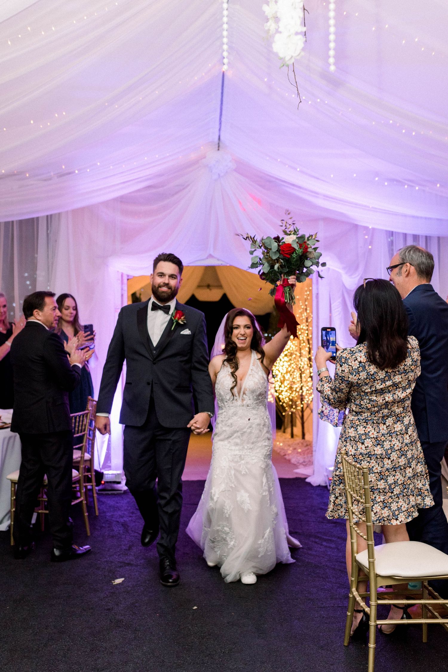 Couple walks down aisle in beautifully decorated wedding reception venue with draped fabric and hanging lights.