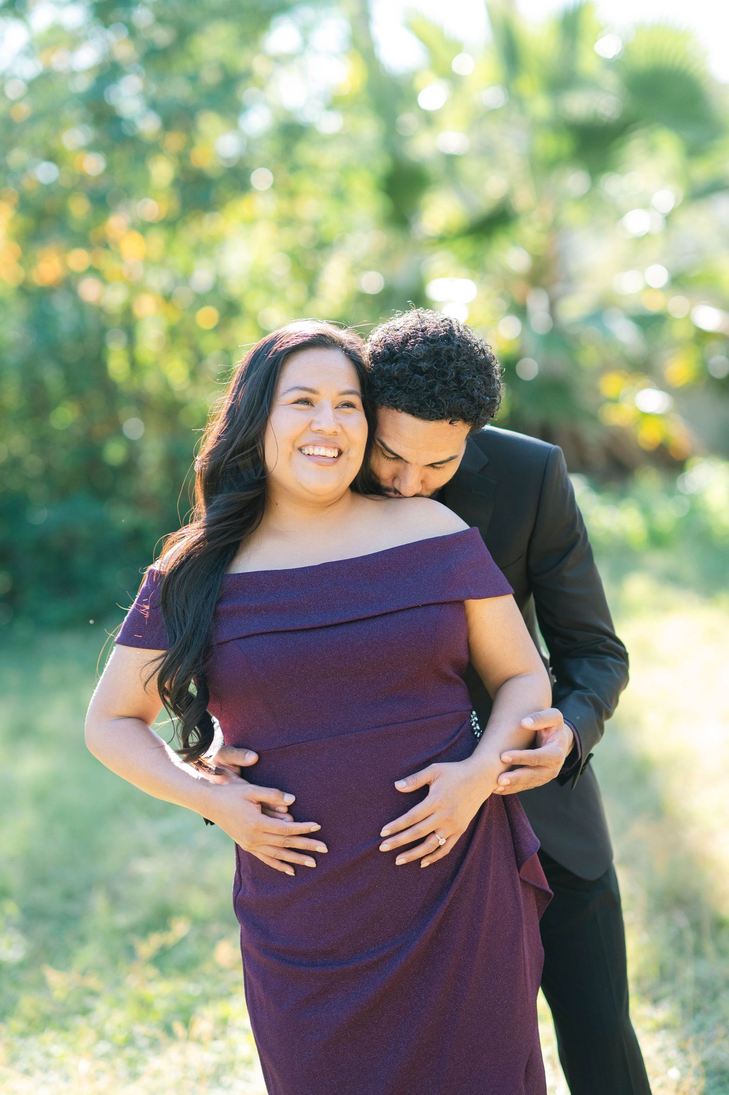 A couple embraces outdoors during a maternity photoshoot, with the expectant mother wearing a purple off-shoulder dress.