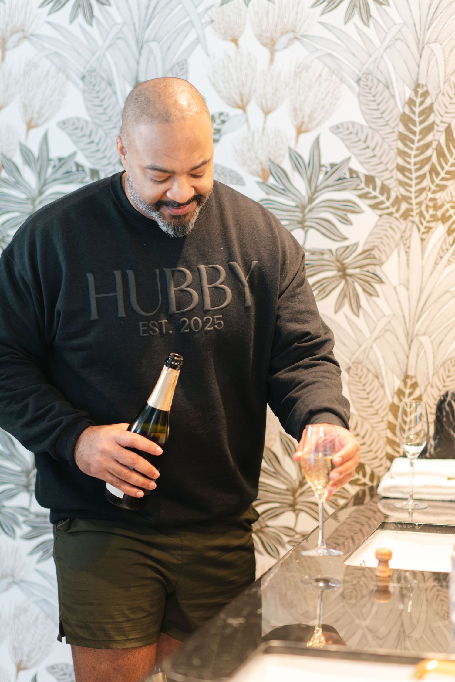 A person in a black 'HUBBY EST. 2023' sweatshirt poses in a series of photos against a floral wallpaper background.