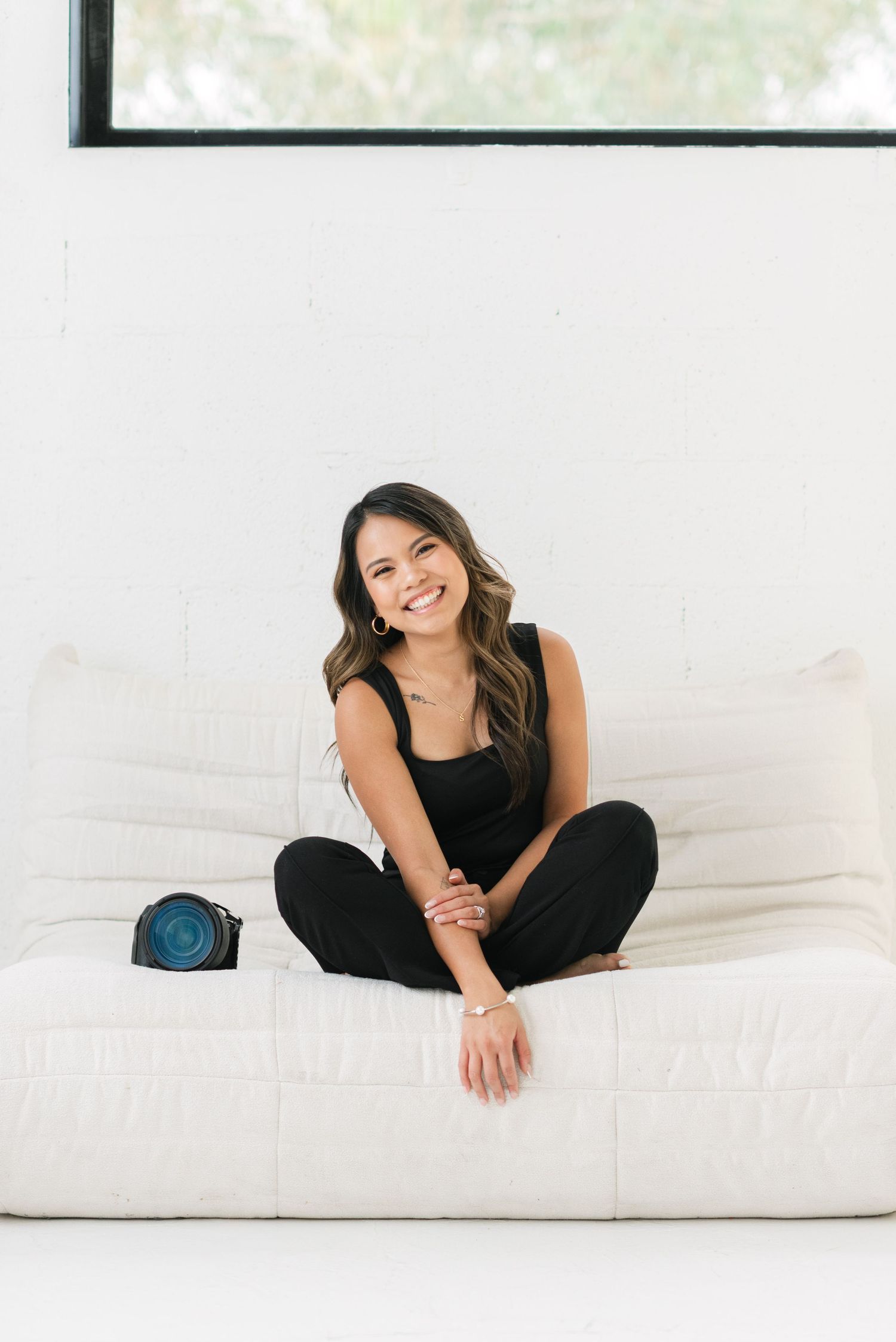 A person wearing black clothing relaxes and smiles while lounging on a white couch in a bright room.