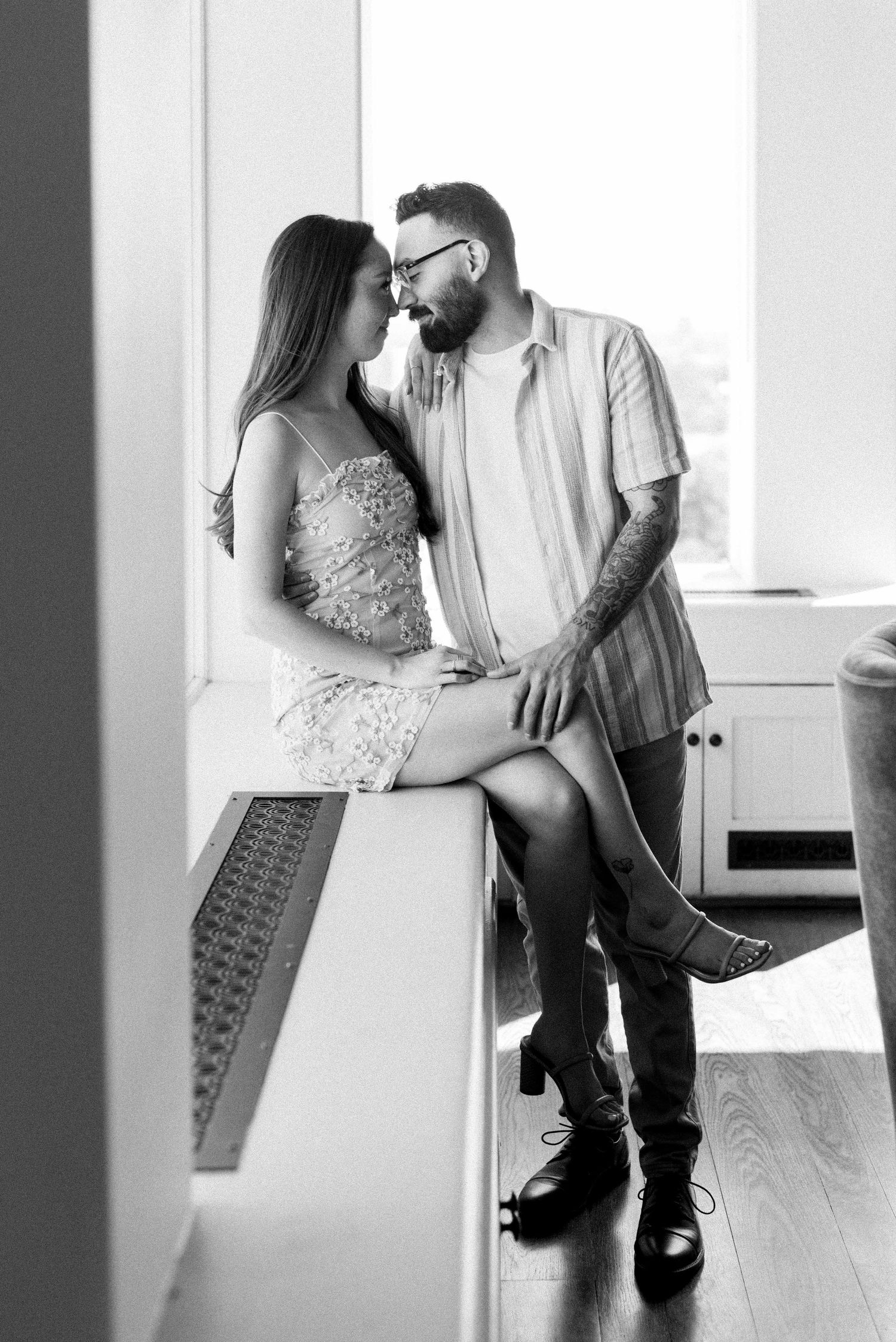 Black and white photo of a couple in casual formal wear sharing an intimate moment by a window.
