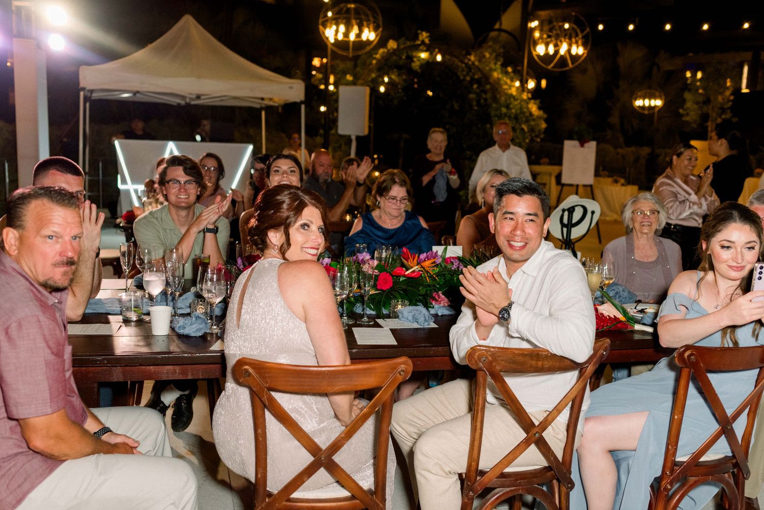 Wedding guests enjoy dinner at outdoor tables decorated with string lights and florals.