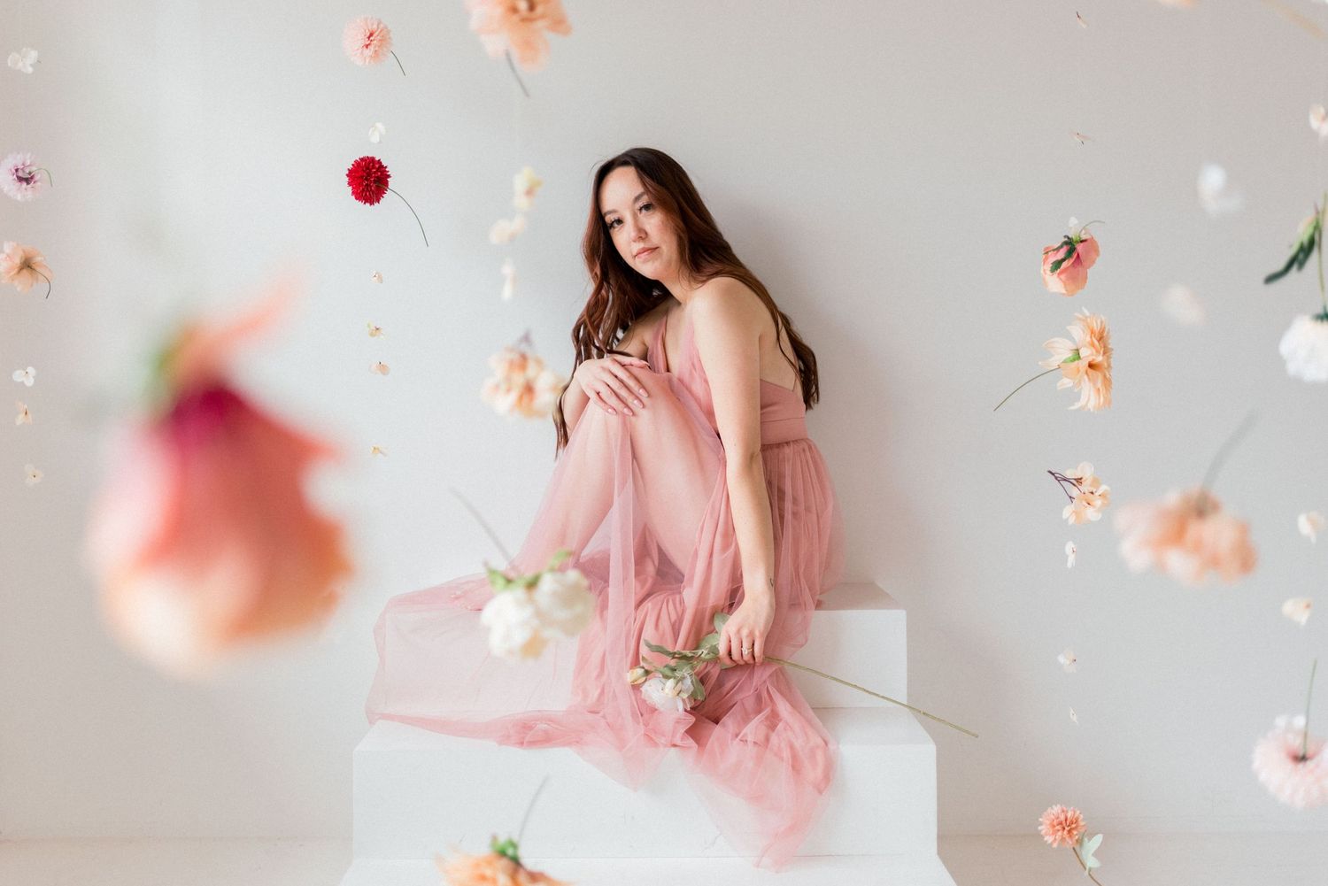 Artistic portrait with floating flowers in pink dress against white backdrop.