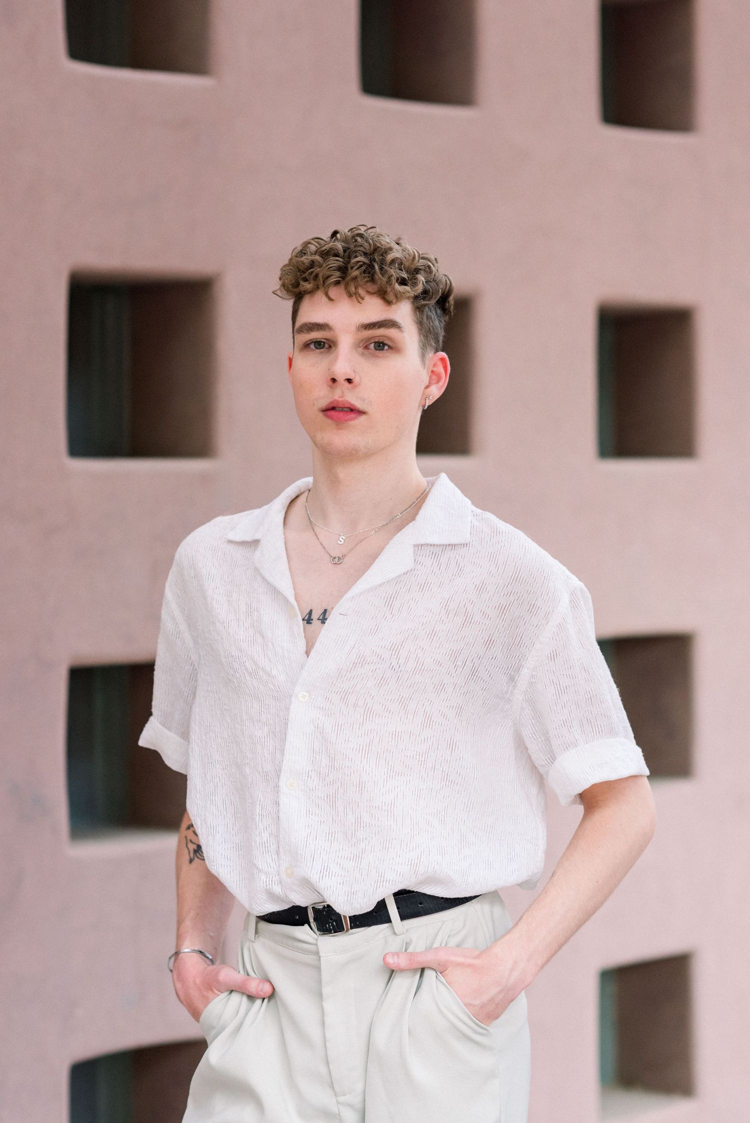Person in white shirt and light pants poses against pink geometric wall.